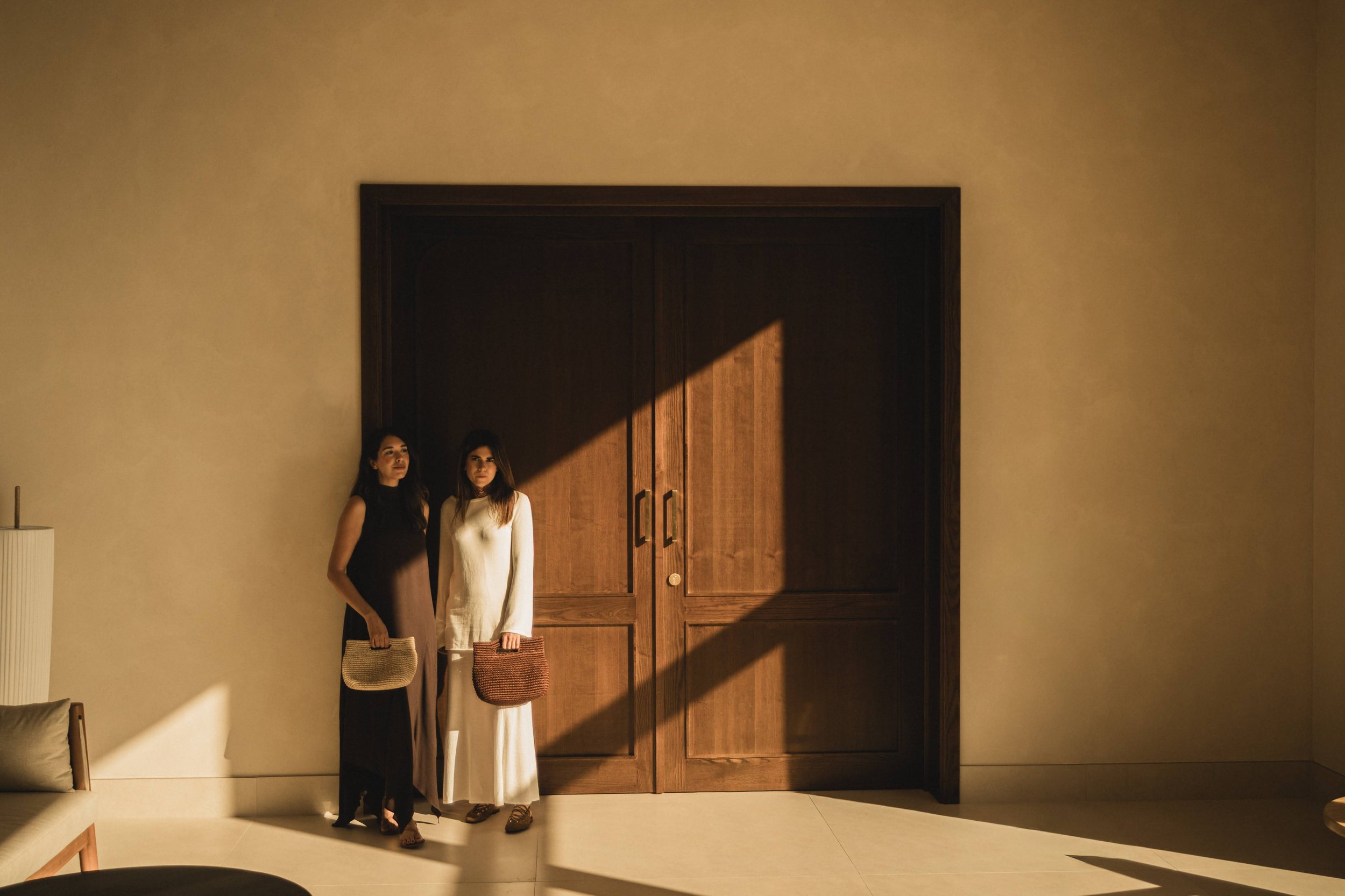 Dos mujeres de pie junto a una puerta de madera en una sala. La mujer a la izquierda lleva un vestido oscuro y sostiene un bolso beige, mientras que la otra lleva un vestido blanco y sostiene un bolso de colores. La luz del sol entra por una ventana, creando una sombra diagonal en la pared.