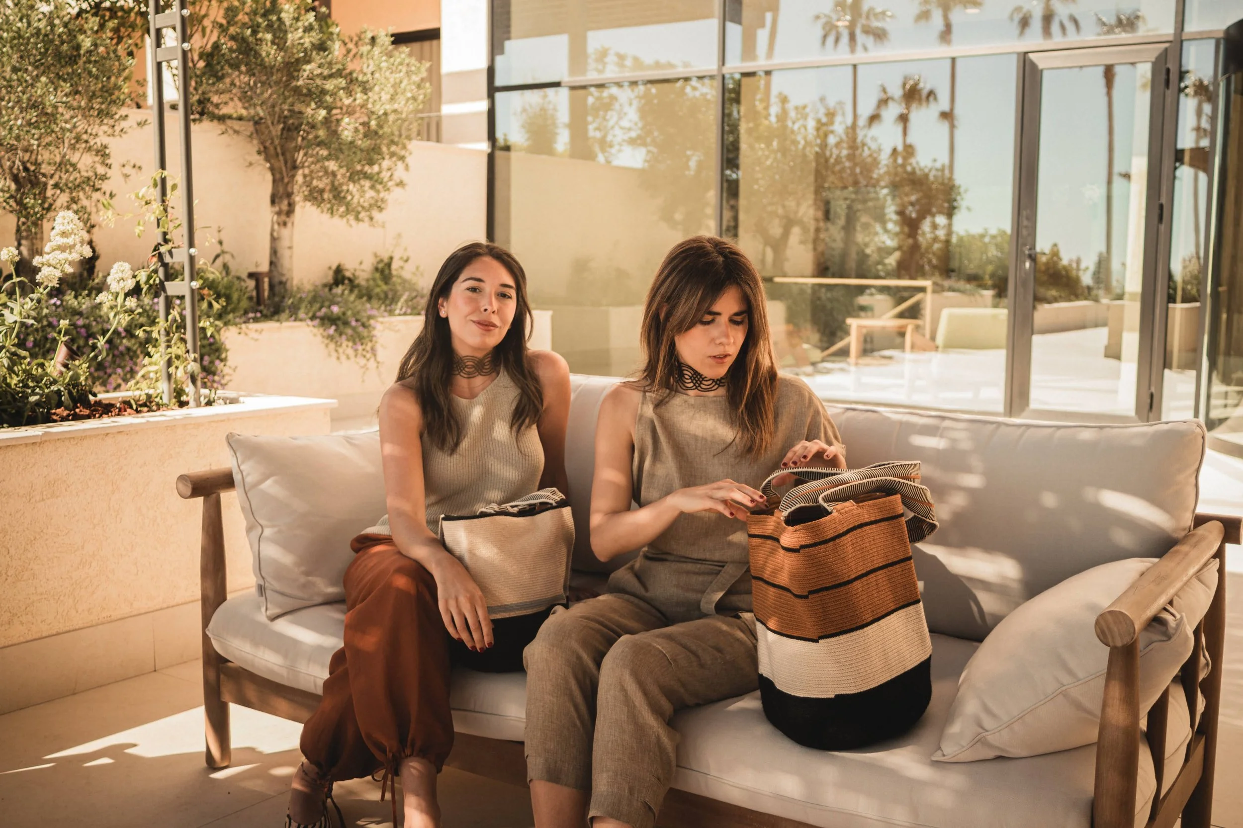 Dos mujeres sentadas en un sillón de mimbre en un espacio cerrado con paredes de vidrio, rodeadas de plantas verdes y flores, una revisando una bolsa y la otra sonriendo.