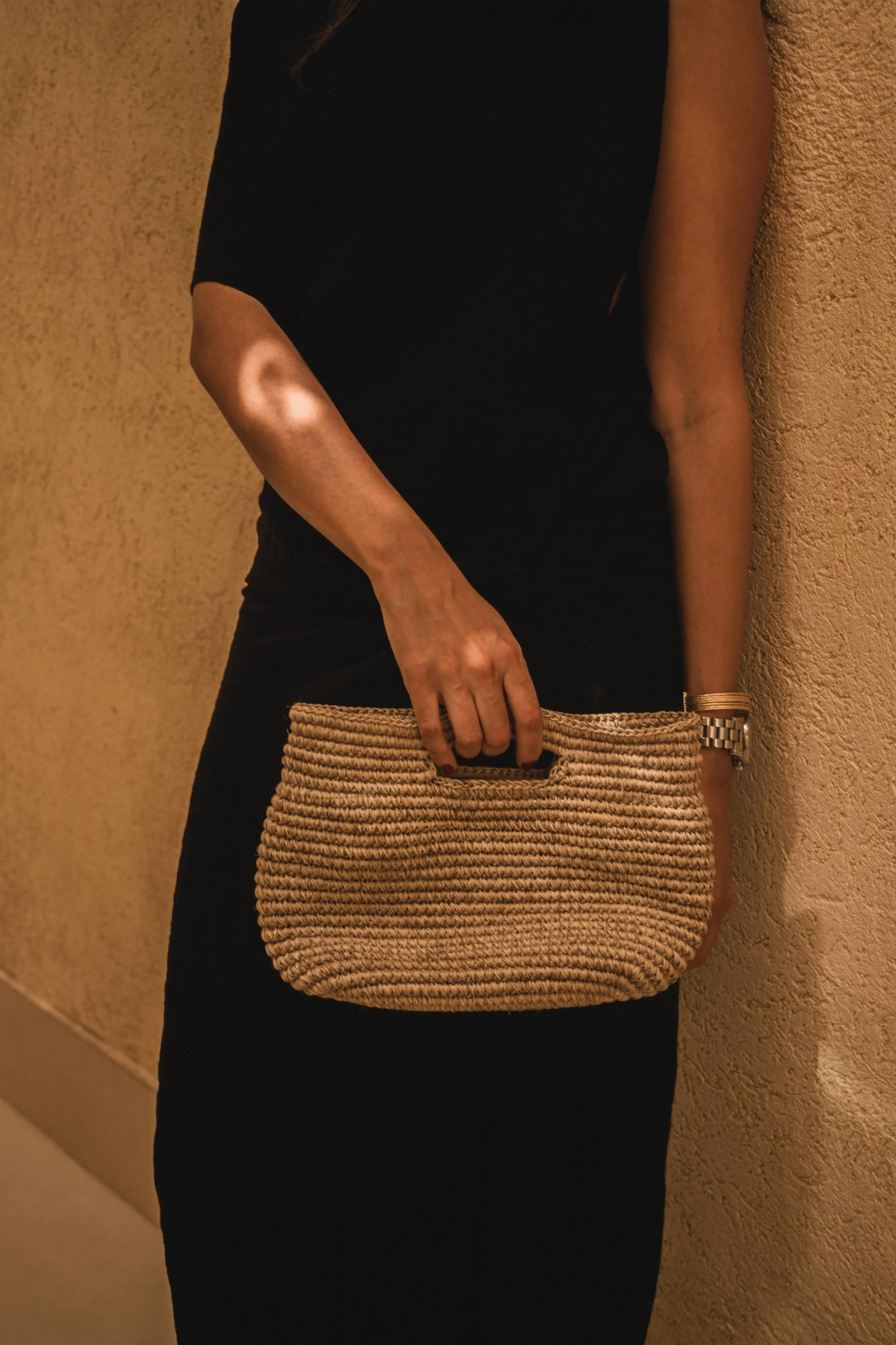 Persona con un vestido negro, sosteniendo un bolso de mano tejido de color beige, usa reloj dorado y tiene el cabello largo.
