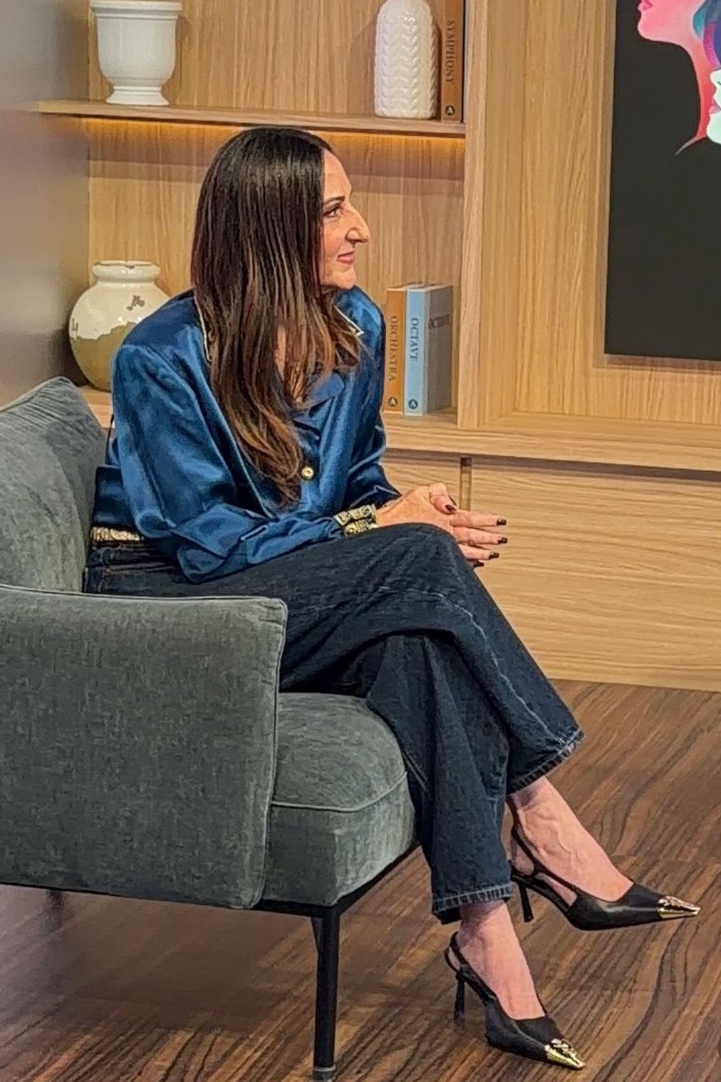 Mujer sentada en un sillón de color gris oscuro en un ambiente moderno, con estanterías de madera detrás, usando blusa de satín azul, pantalones vaqueros y zapatos de tacón negro con detalles dorados.