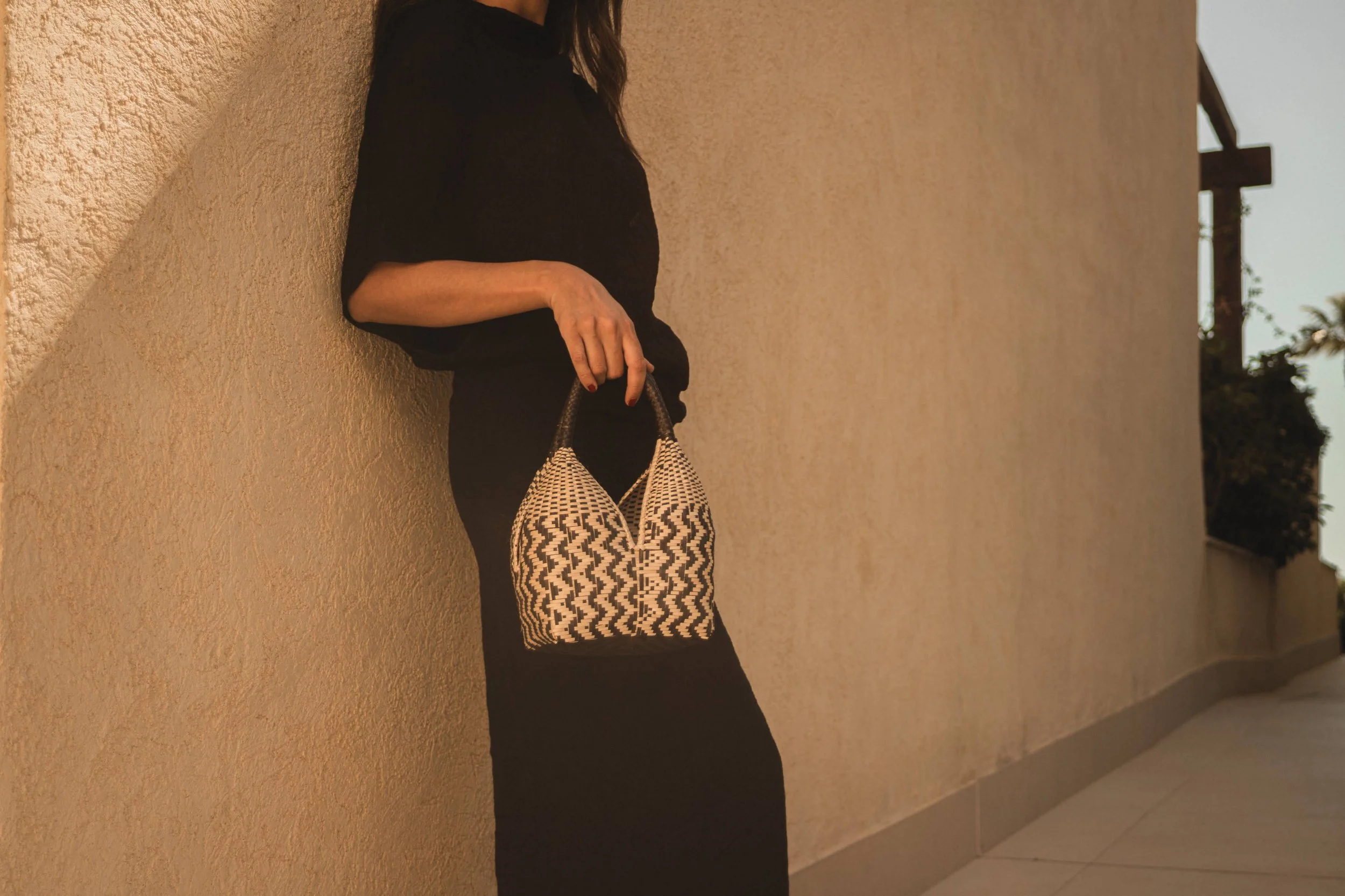 Persona de espaldas apoyada en una pared de color beige, usando ropa negra y sosteniendo una bolsa con diseño de patrones en blanco y negro, en un entorno exterior con vegetación y estructura de fondo.
