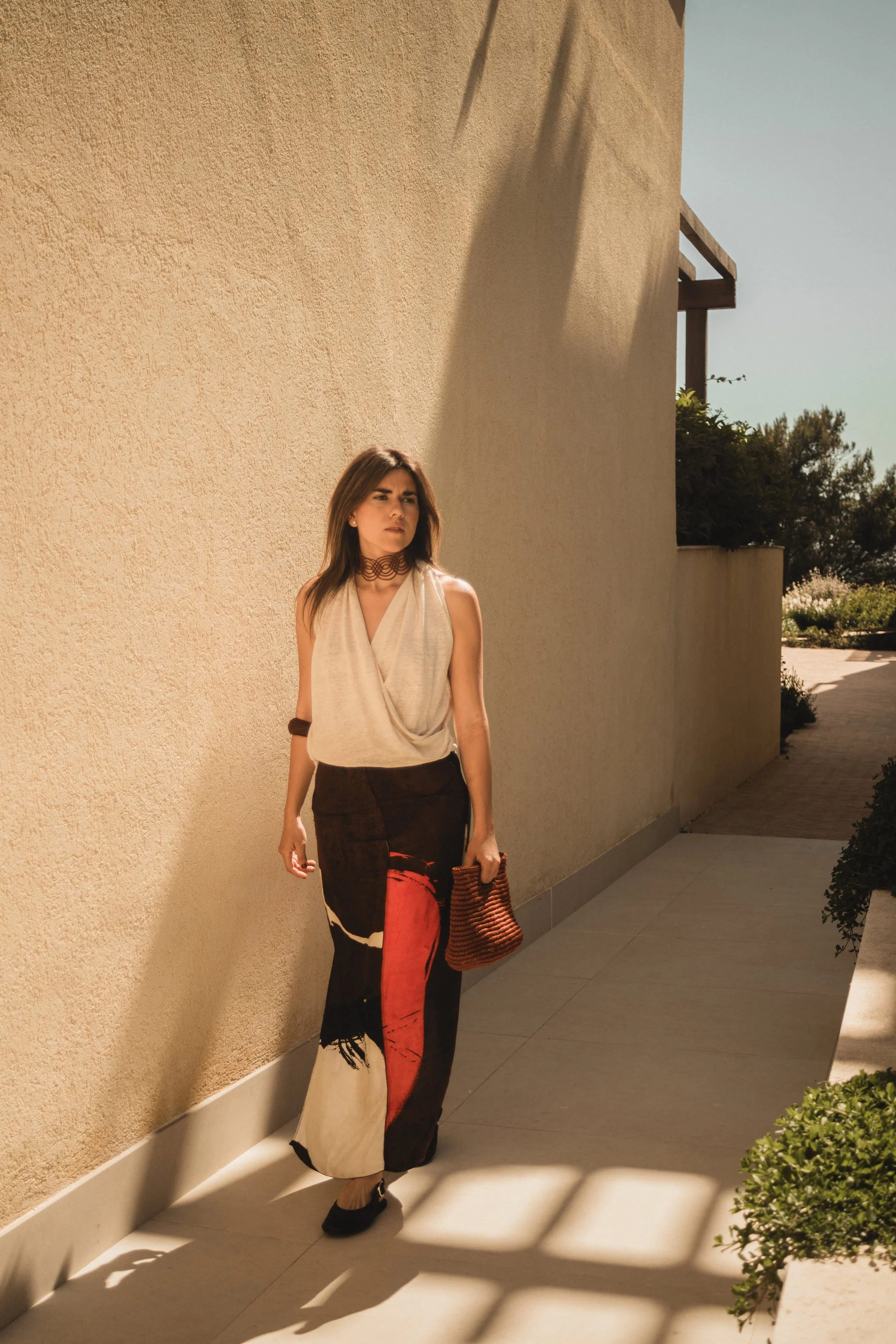Mujer con vestido beige y pantalón largo con diseño abstracto en tonos negros, rojos y beige, sosteniendo una bolsa de mano marrón, caminando al aire libre en un día soleado.