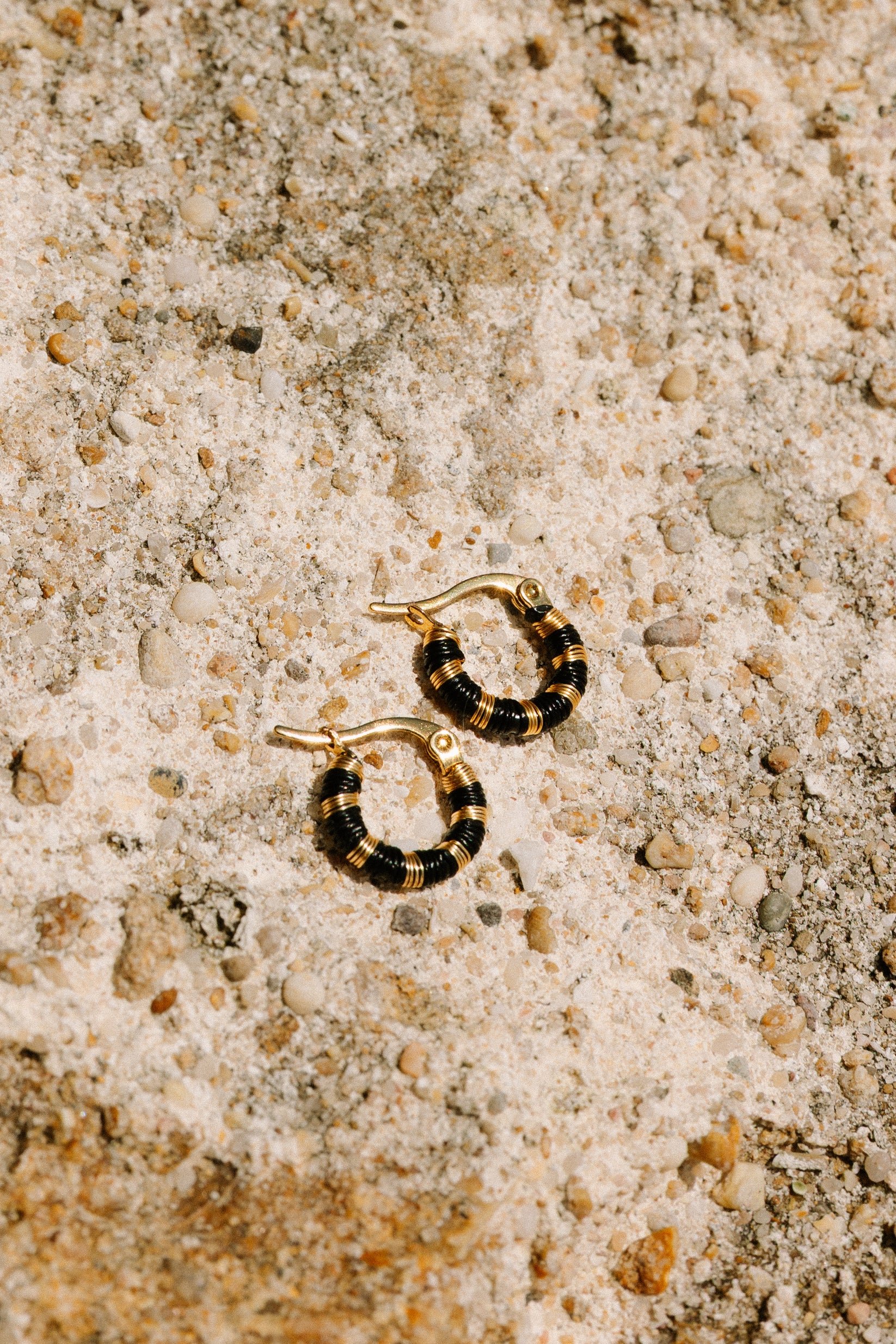 Aretes en forma de aro con cuentas negras y doradas sobre arena con pequeñas conchas y piedras.