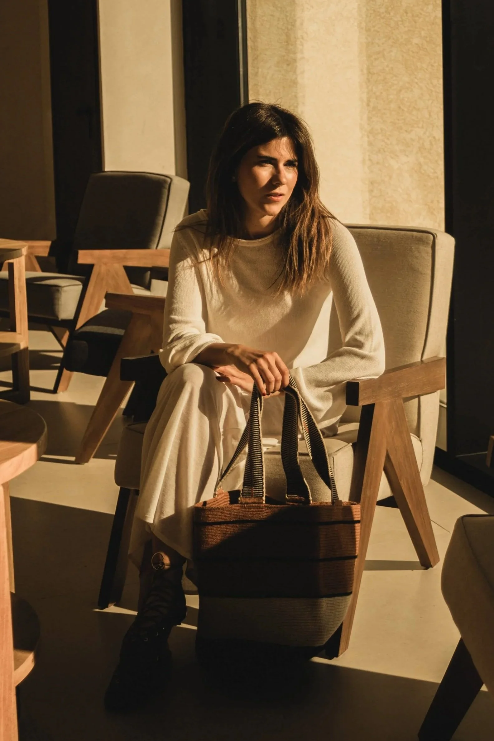 Mujer sentada en una silla de madera en un ambiente con luz tenue, lleva pantalones blancos y una camiseta beige, y sostiene un bolso de mano. La escena parece ser en un lugar moderno y elegante.
