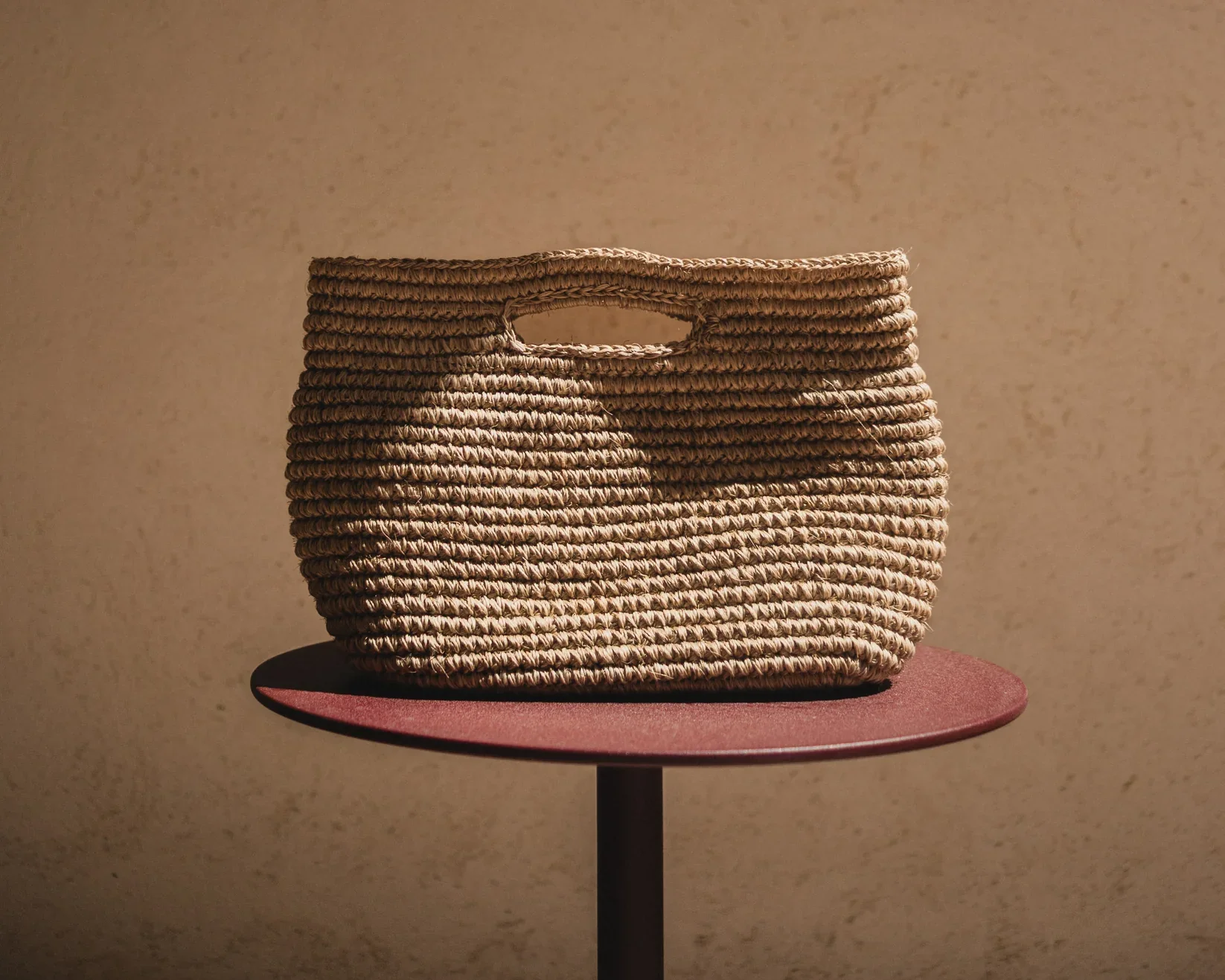 Bolsa de mimbre con doble asa, colocada sobre una mesa de color rosado oscuro, con fondo de pared beige.