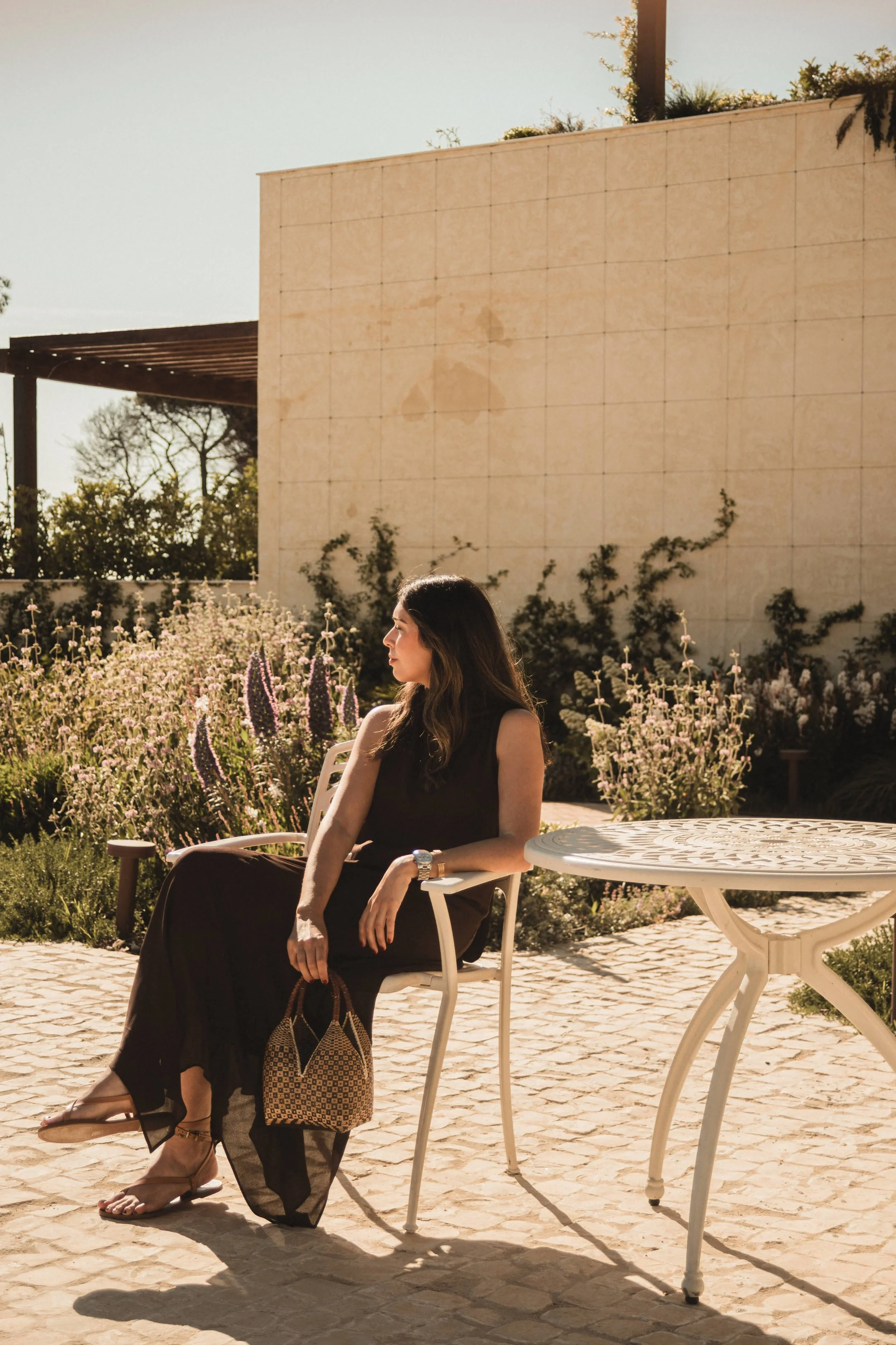 Una mujer sentada en una silla de jardín en un patio rodeado de flores y plantas, con una pared y un árbol en el fondo, en un día soleado y cálido.