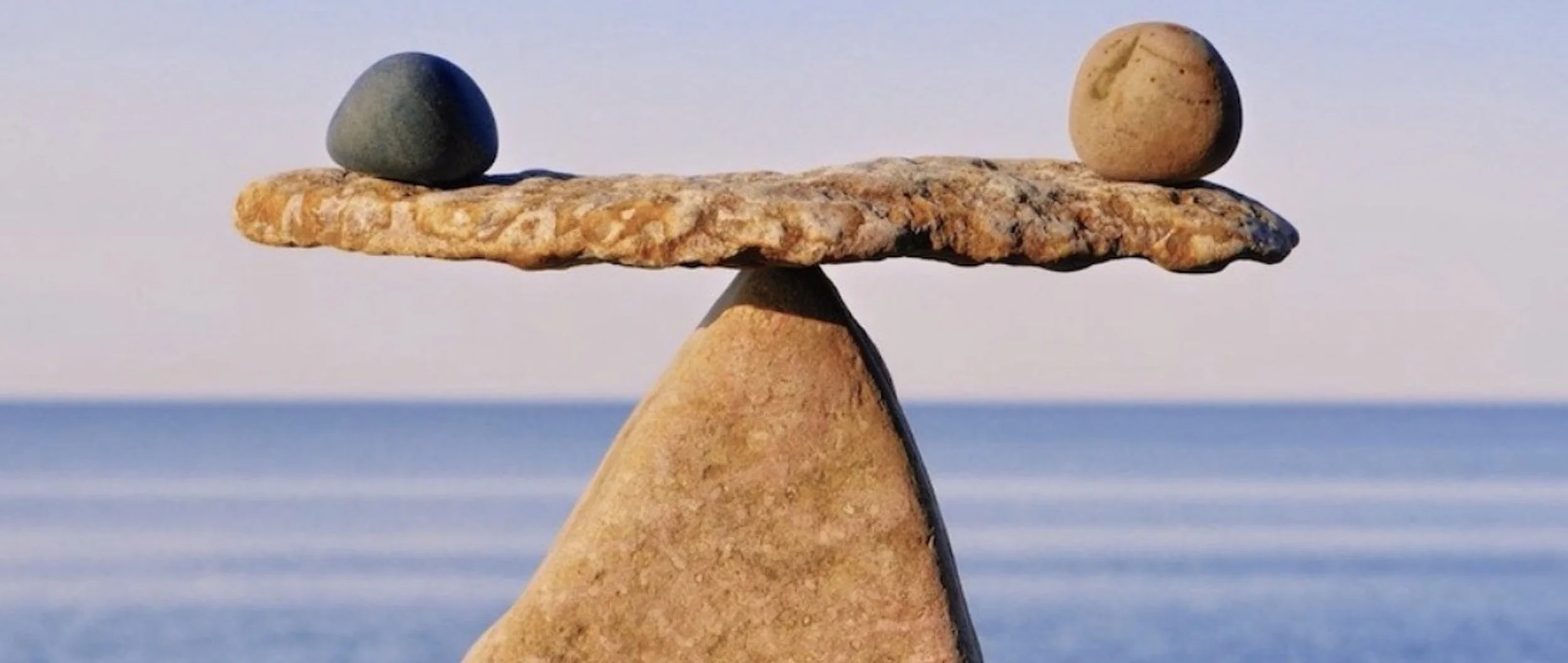 A balanced stone sculpture with two rocks on a flat stone on top of a cone-shaped stone, creating a seesaw effect at the beach with the ocean in the background.