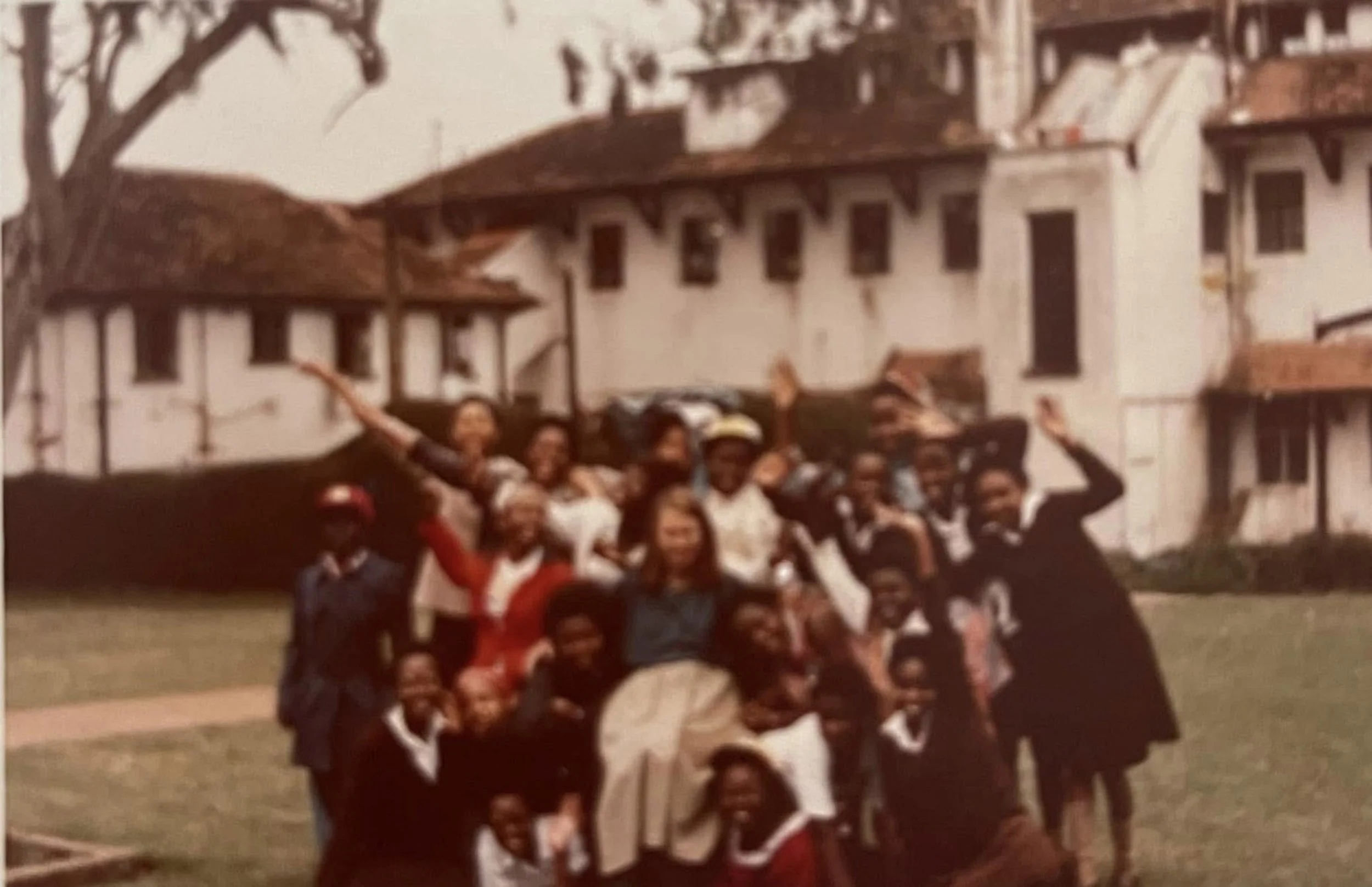 A group of children and adults posing outdoors in front of a building with books on a balcony, some children wearing sports jerseys, and adults standing behind the kids, appearing to celebrate or mark an event.