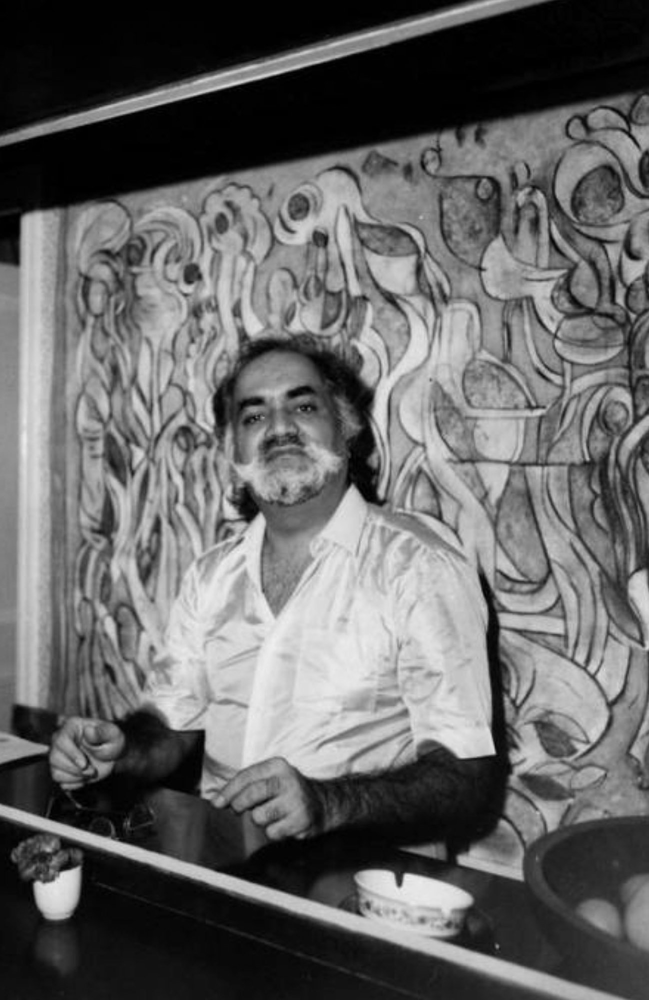 A man with dark hair and a beard sitting at a table with coffee mugs, in front of a colorful mural with abstract designs.