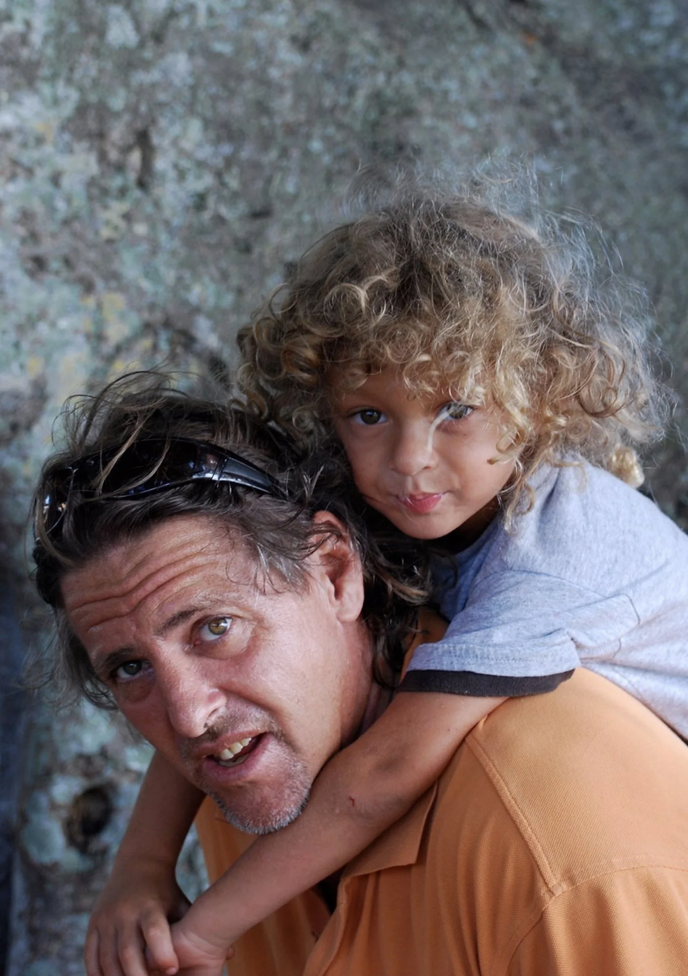 A man with gray hair and a beard giving a piggyback ride to a young girl with curly blonde hair and brown eyes, both outdoors against a blurred natural background.
