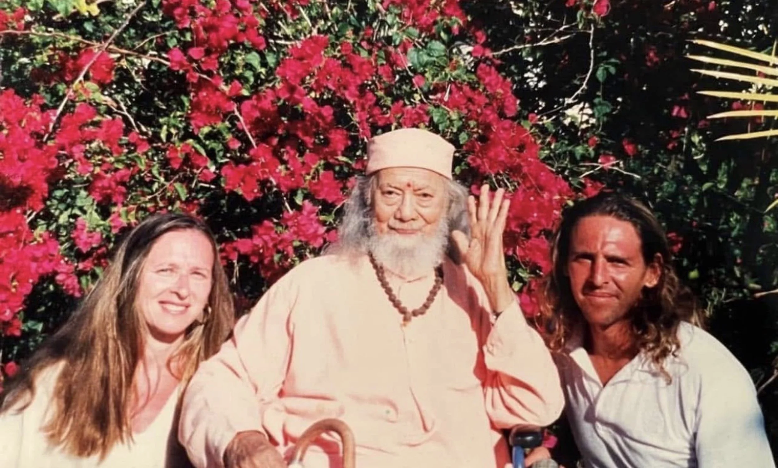 Three people standing in front of a large, bright red flowering bush. The person in the center is an elderly man with long white hair and a beard, wearing a light pink robe, a beige hat, and multiple beaded necklaces, and is making a gesture with his