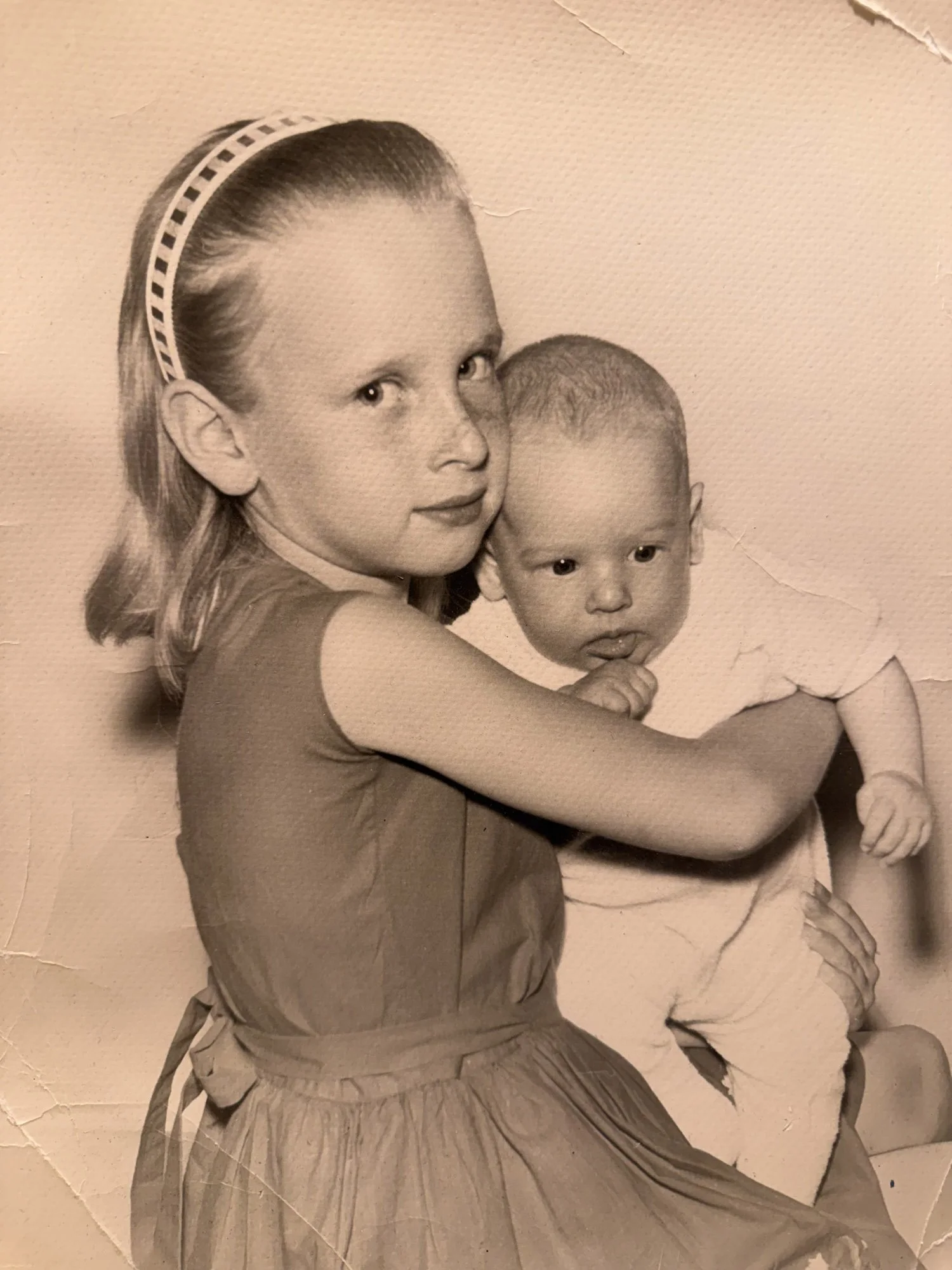 Black and white photo of a young girl holding a baby boy, both facing camera, with torn and wrinkled paper texture background.