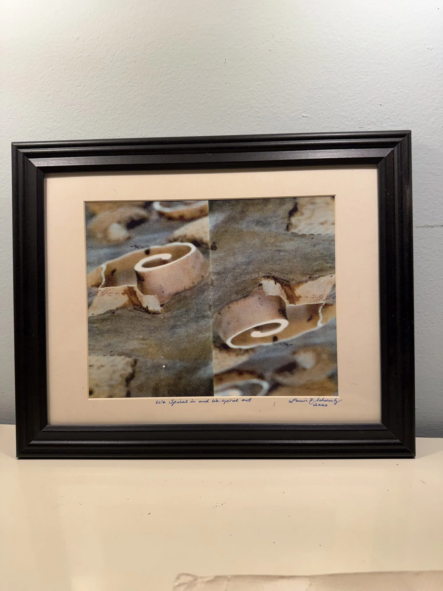 Framed artwork of a spiral shell on a rocky beach, with handwritten text at the bottom reading 'We spiral in and we spiral out - Laurie T. Scholz 2020'.