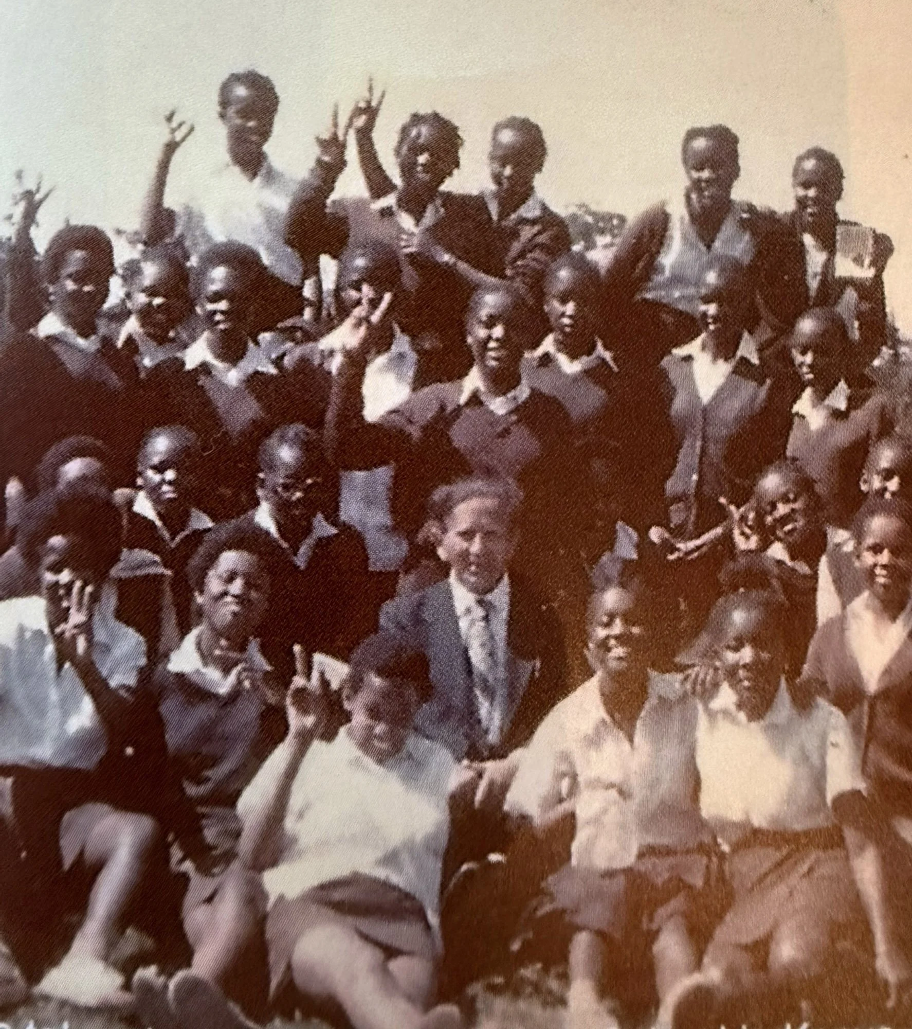 Black and white photograph of a group of children and a few adults, with some standing and others sitting on the ground, outside on a sunny day.