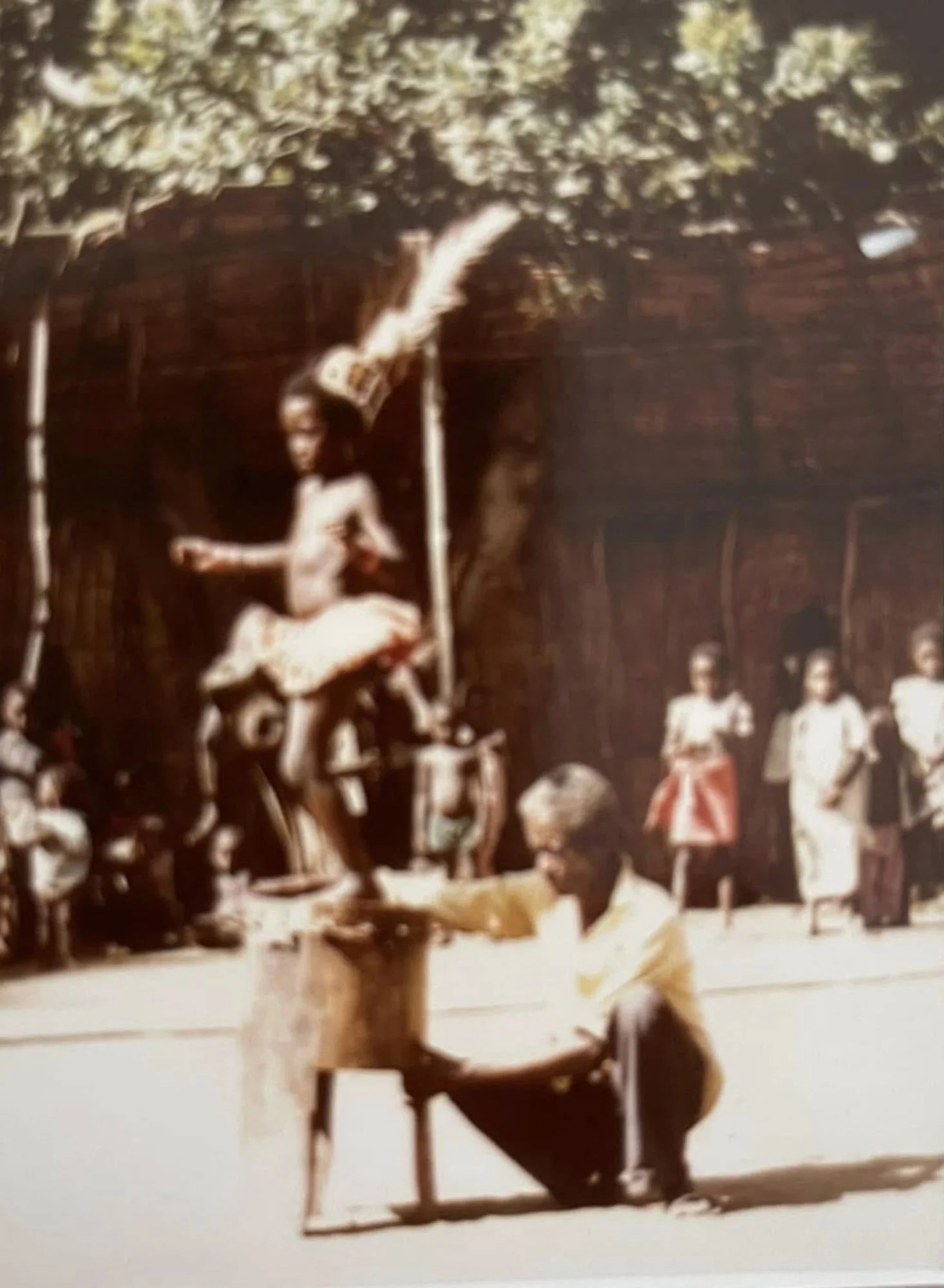 A young girl sits on a bench playing a piano outdoors, with other children in the background and trees overhead.