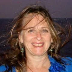 Close-up of a smiling woman with light brown hair, wearing turquoise earrings and a blue top, outdoors against a cloudy sky.