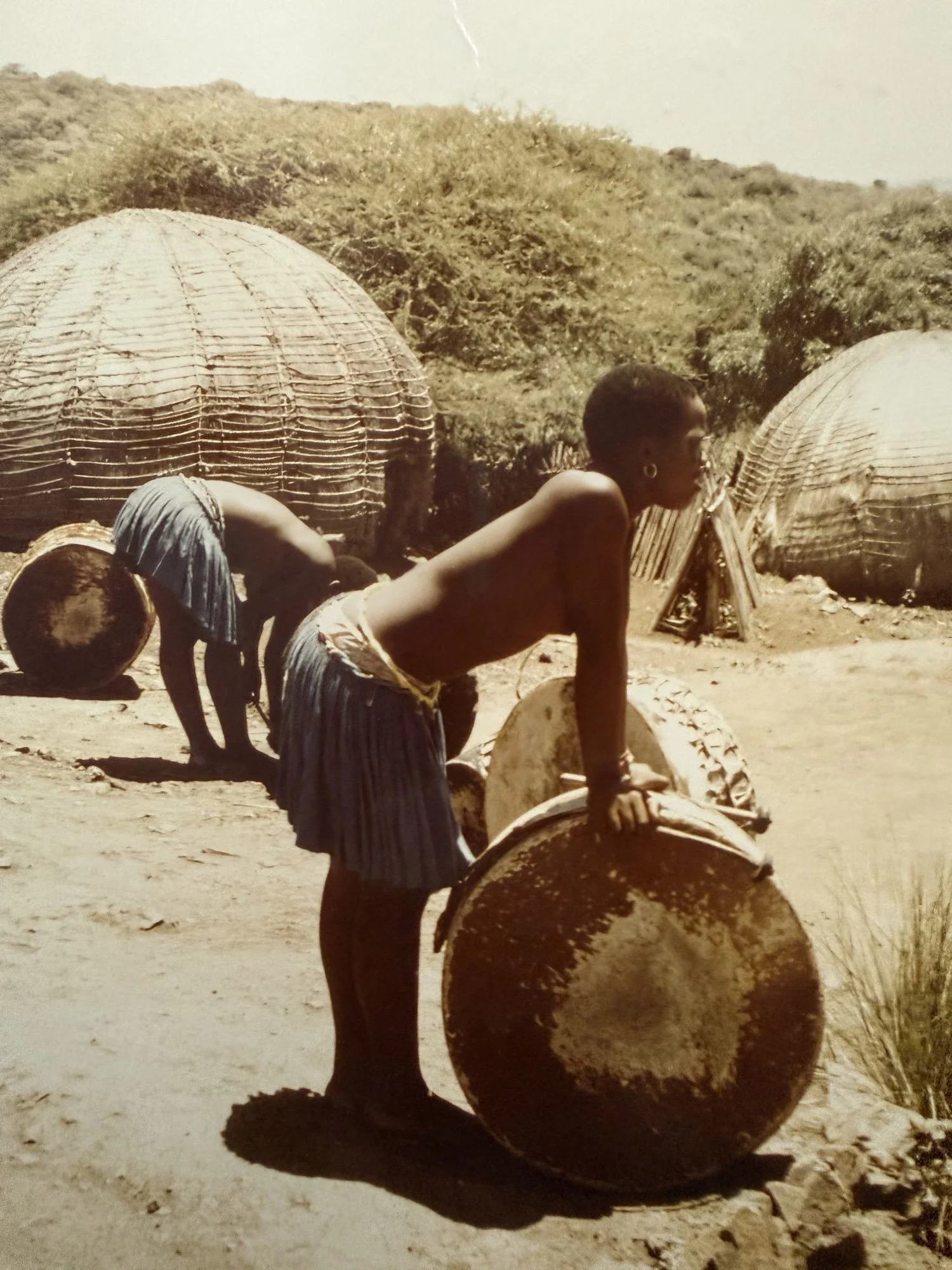 Children playing with large clay pots outdoors in a rural setting, with grass huts and hilly terrain in the background.