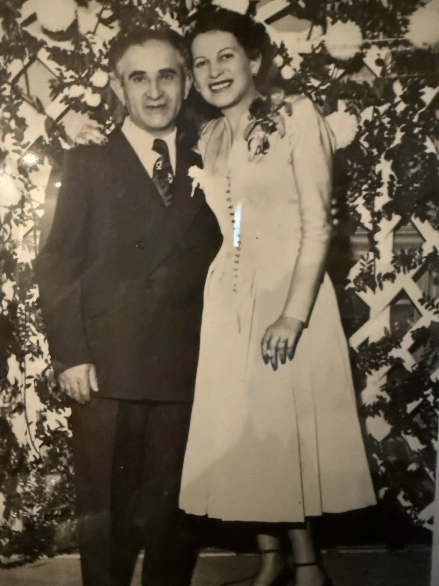 Black and white photo of a man and woman standing closely together outdoors, with the woman's arm around the man's shoulder, in front of a plant with large leaves.
