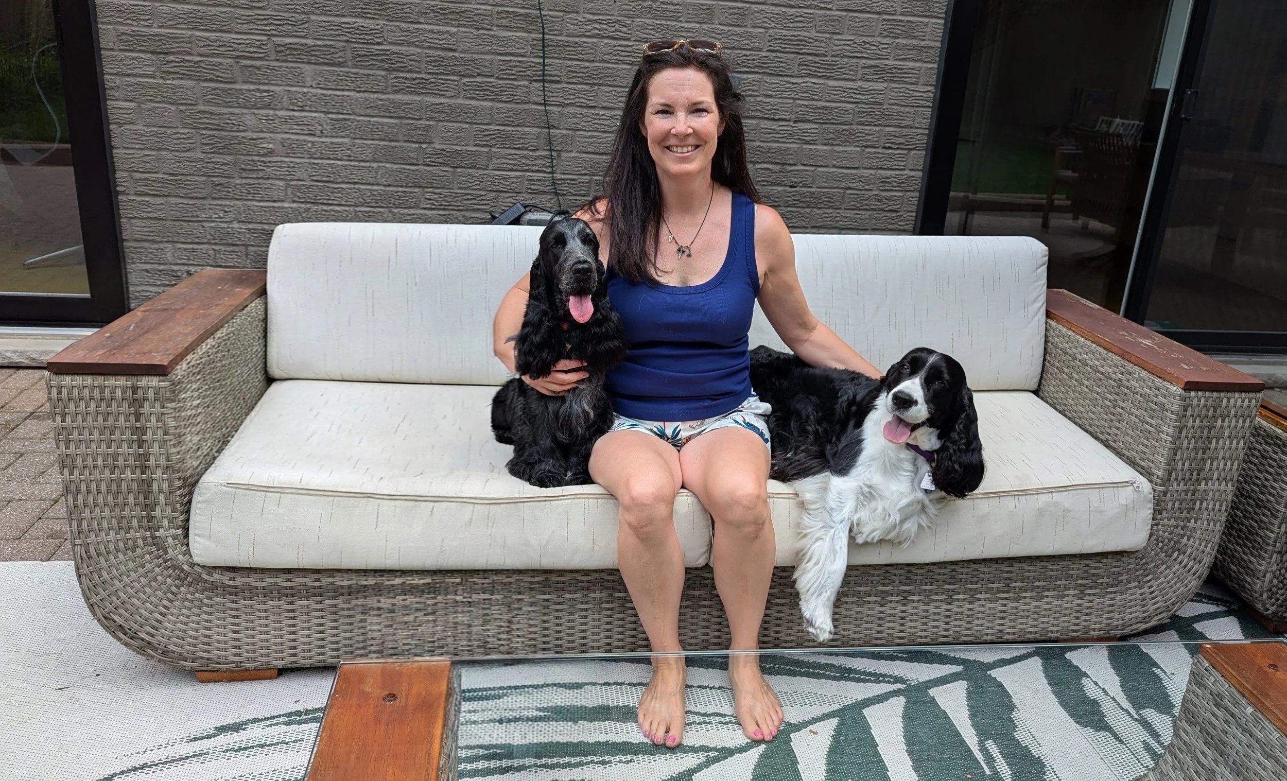 A woman sitting on an outdoor sofa with two spaniels, one of which is a blue roan English Cocker Spaniel, smiling at the camera.