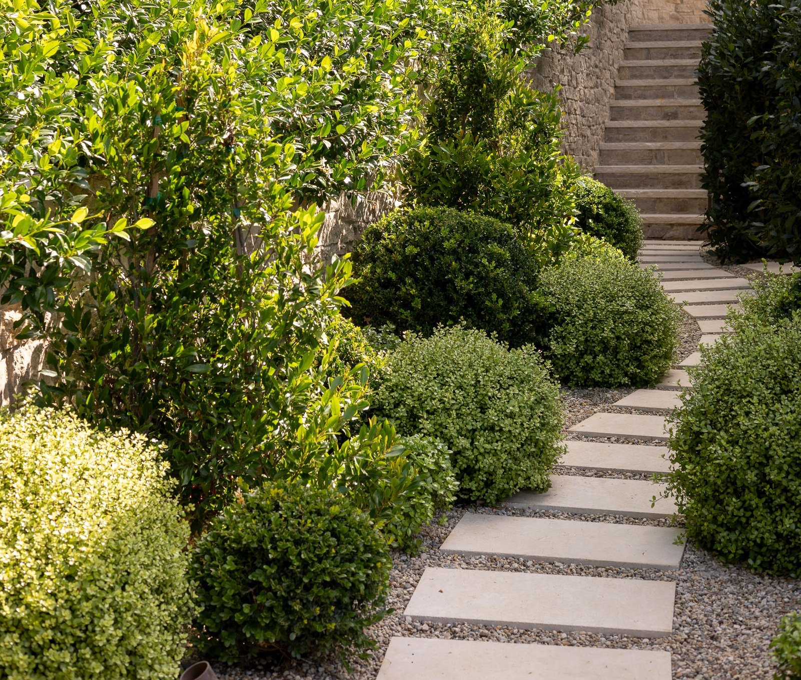 A stone pathway with rectangular stepping stones surrounded by green bushes and plants, with stairs in the background leading up along a stone wall.