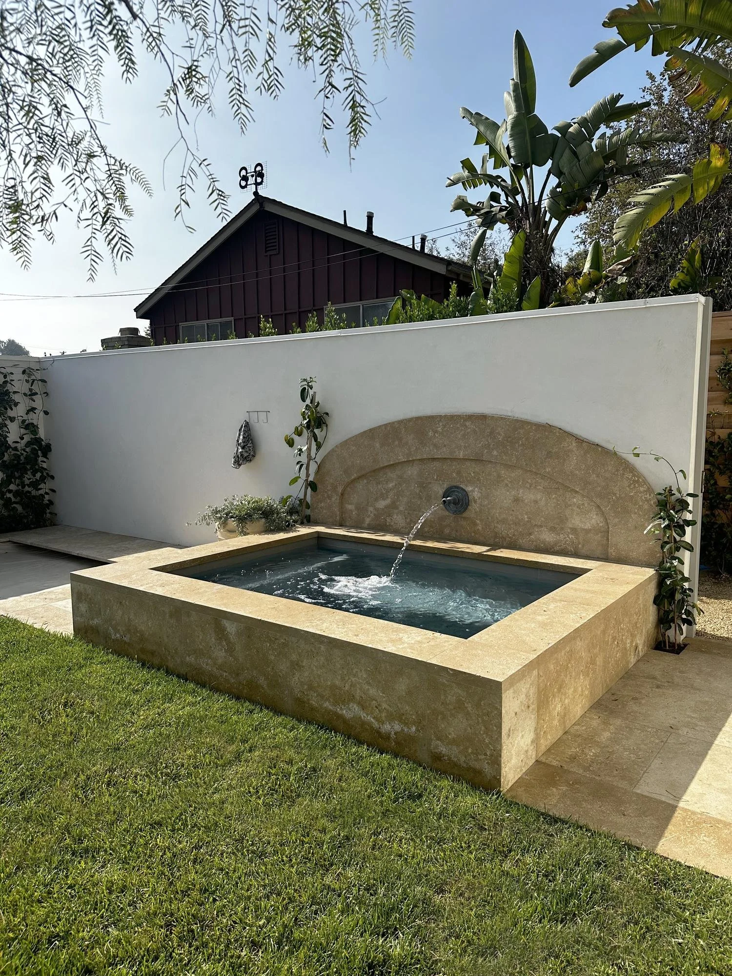 Backyard scene with a rectangular stone fountain with water spout, green grass in the foreground, white privacy wall, and lush tropical plants behind it, with a dark-colored house and blue sky in the background.