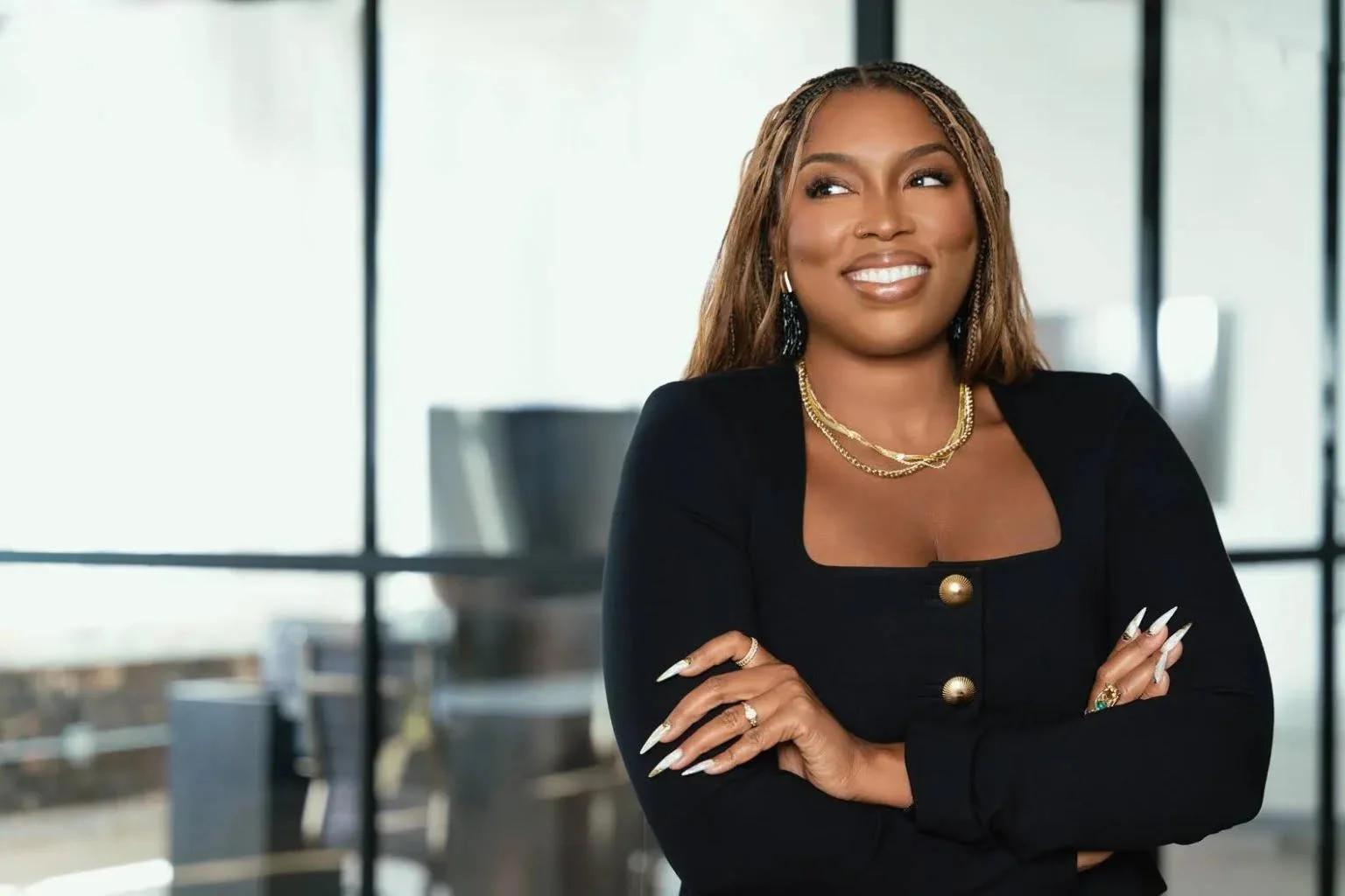 A confident woman with braided hair and brown skin, wearing a black blazer with gold buttons, jewelry including layered necklaces, earrings, and rings, standing in a modern office with glass walls and a city view.