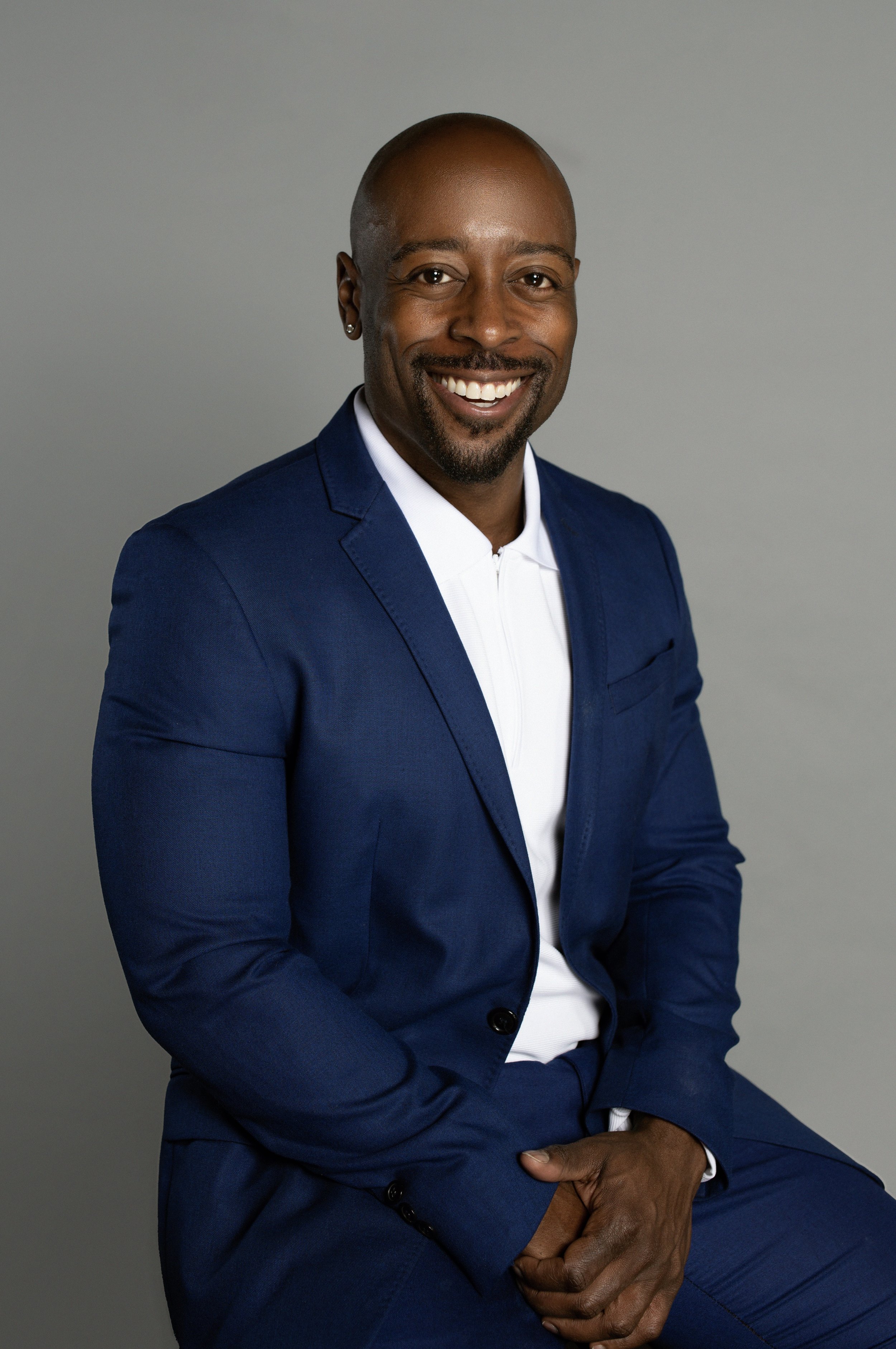 Headshot of a smiling man wearing a blue suit and white shirt, seated against a plain gray background.