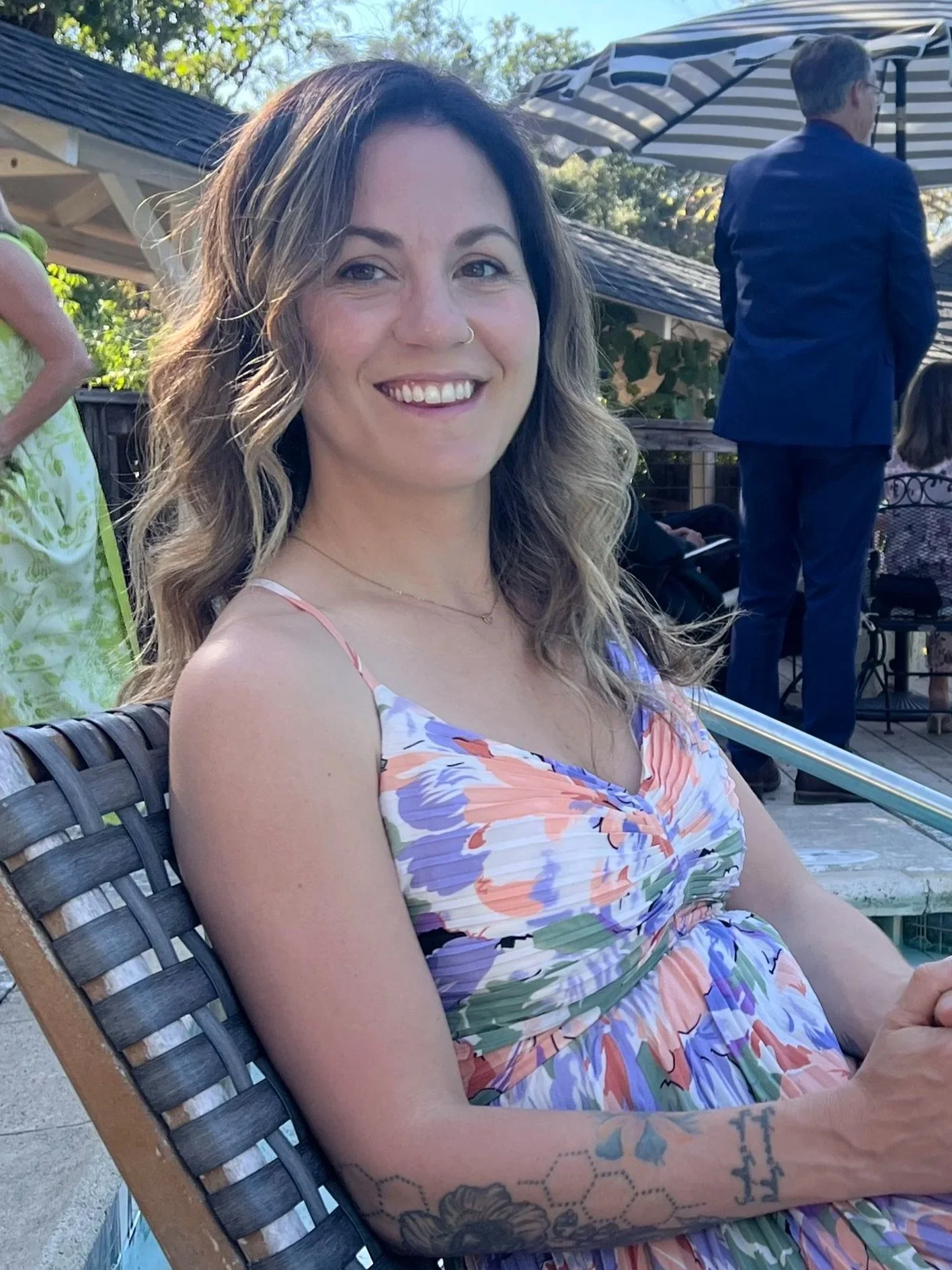 A young woman with wavy hair and a nose ring smiling, sitting outdoors on a wooden chair wearing a colorful floral dress. In the background, there are people, a striped umbrella, and trees.