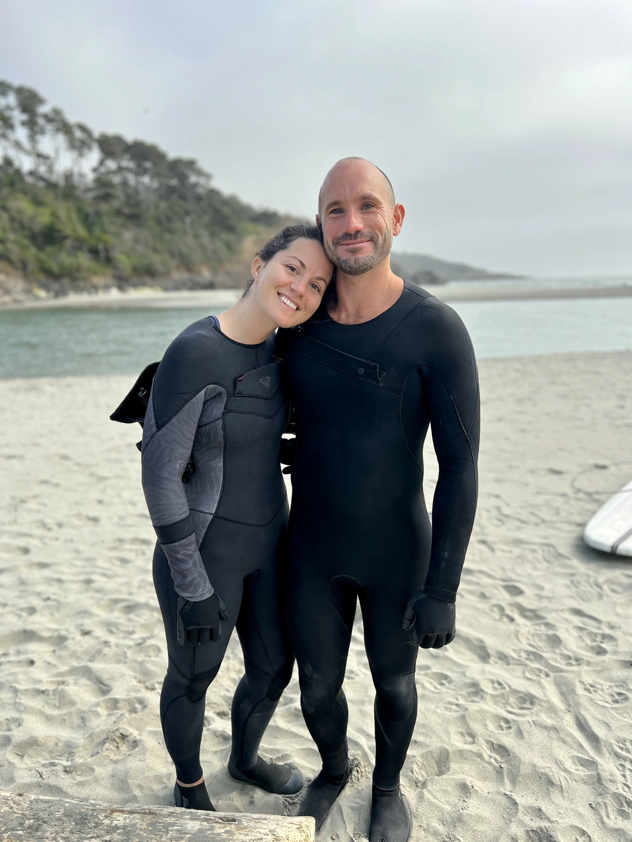 A smiling woman and a serious man in wetsuits standing close together on a beach, with trees and the ocean in the background.
