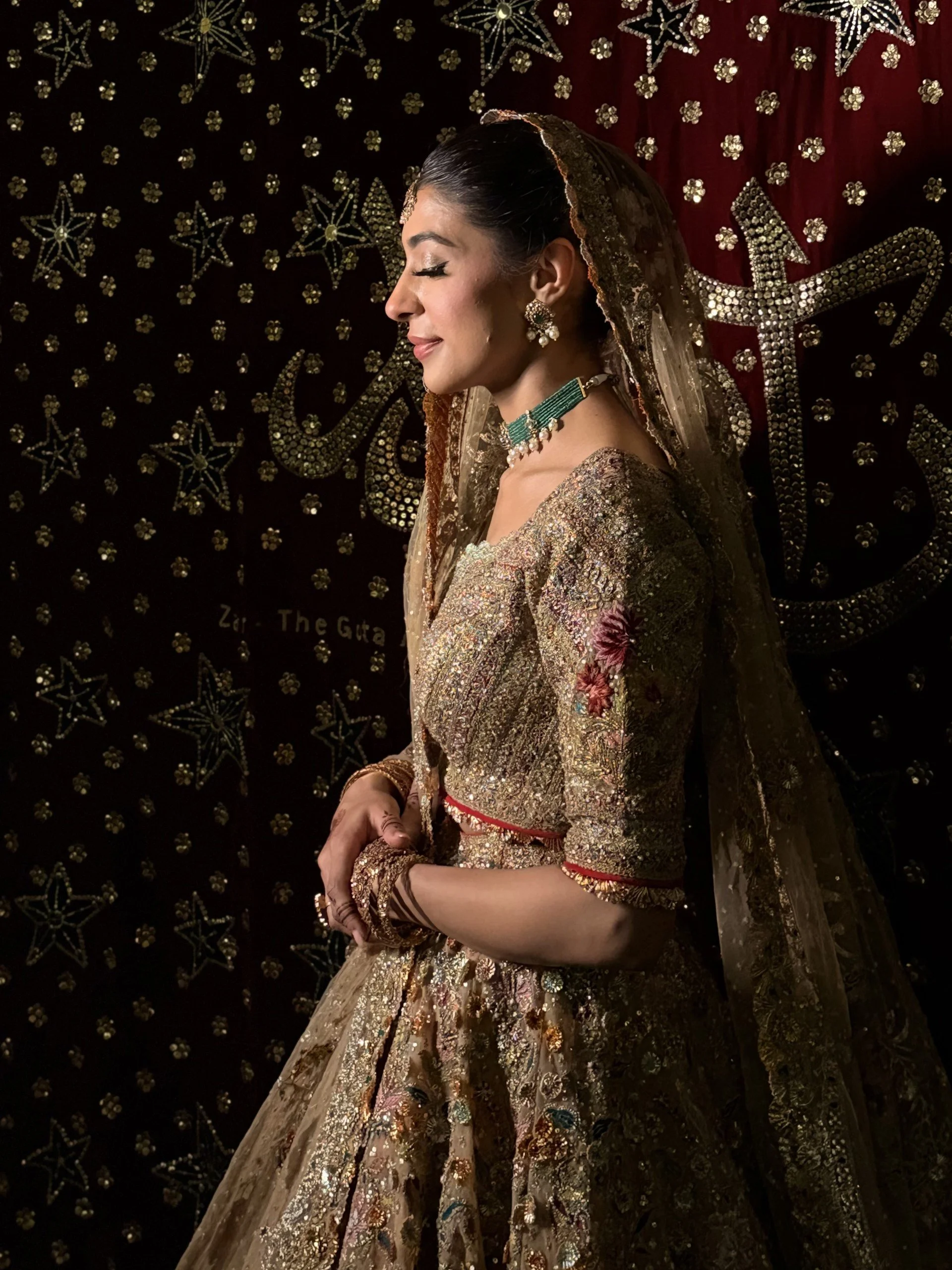 A woman in traditional Indian attire standing with her eyes closed, wearing a heavily embroidered gold and pink sari with jewelry including earrings, a necklace, bangles, and a head covering, against a decorative background with star and flower patterns.