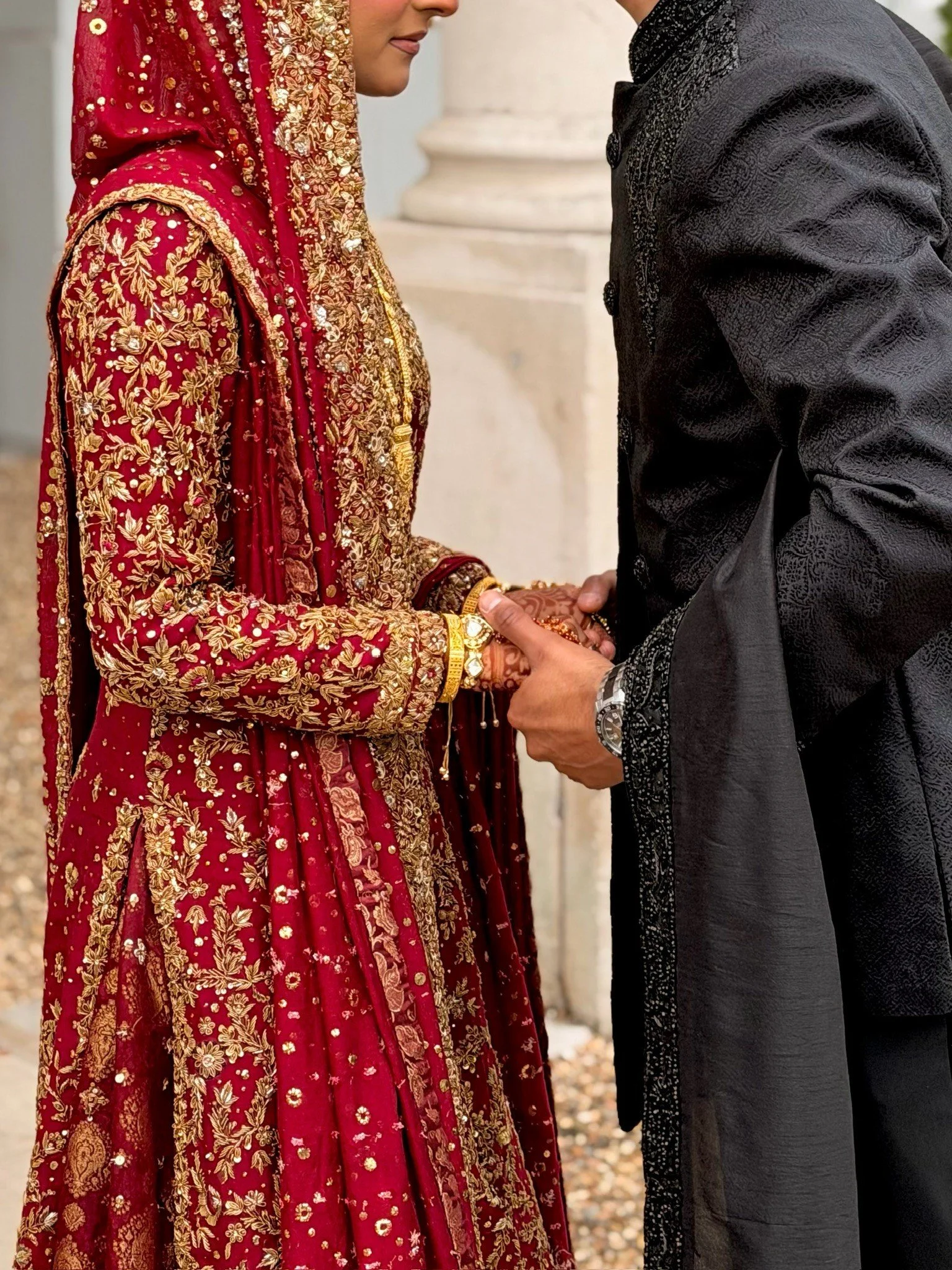 A close-up of a bride and groom holding hands during a wedding ceremony, with the bride wearing a red and gold embroidered traditional dress and the groom in a black textured suit.
