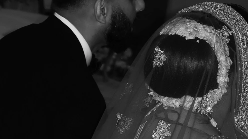 A man and woman dressed in formal wedding attire, with the woman wearing a lace wedding dress and veil, standing close together during a wedding ceremony.
