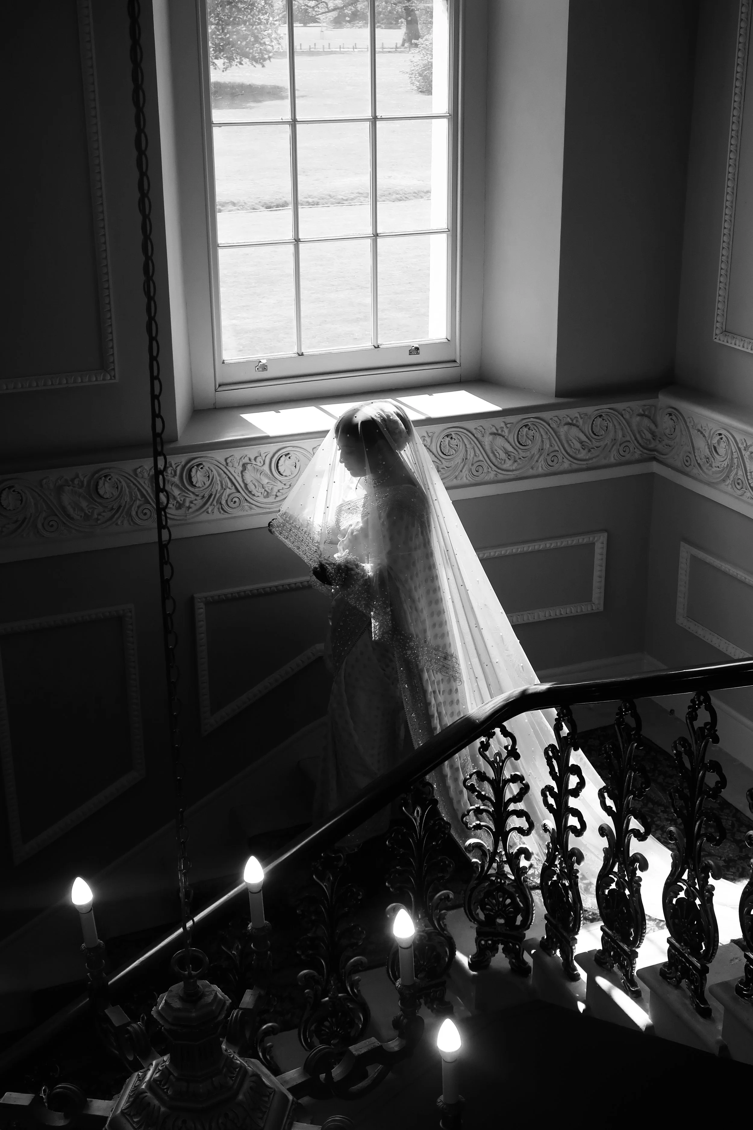 Black and white photo showing a staircase with an ornamental wrought iron railing, a chandelier with a veil draped over it, and a large window in the background revealing an outdoor scene with grass, trees, and a fence.