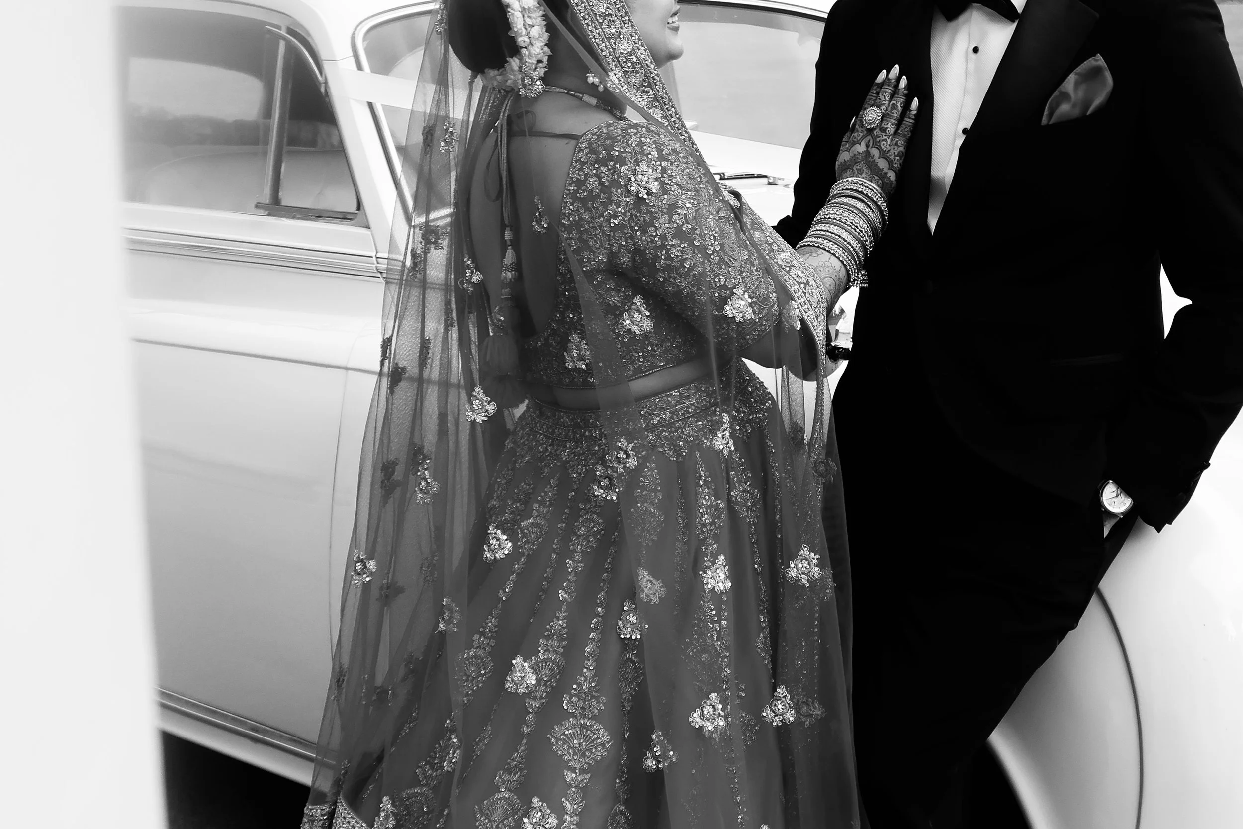 A woman dressed in traditional Indian bridal attire standing close to a man in a tuxedo, near a car.