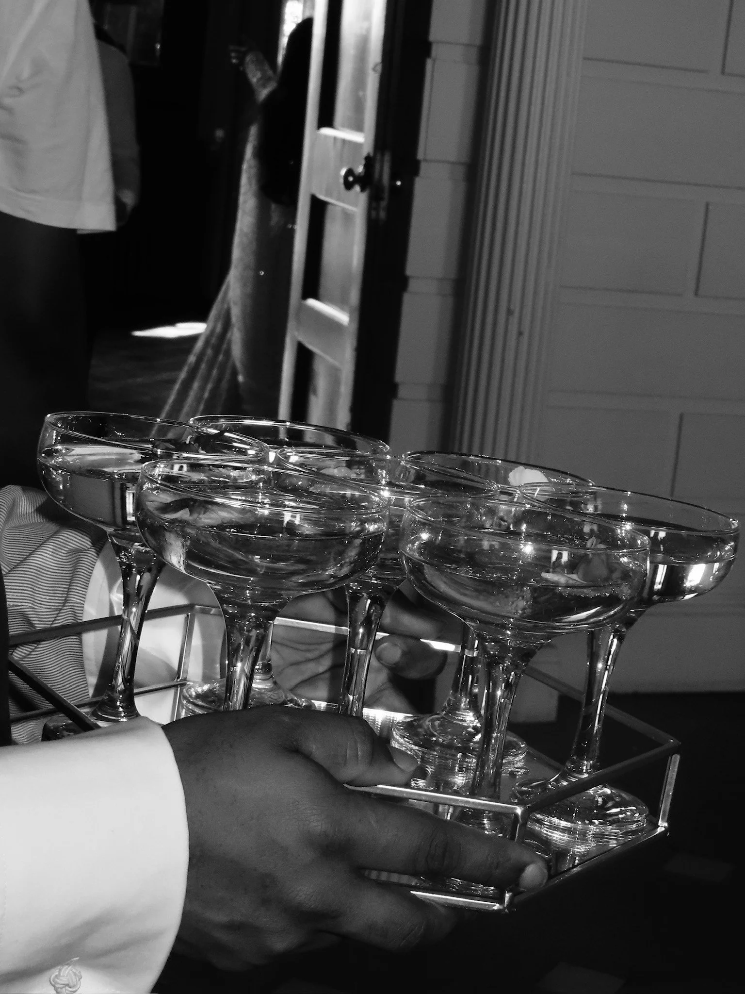 Multiple empty champagne glasses on a silver tray held by a person with a visible hand and arm. Background shows a wooden door and an interior space.