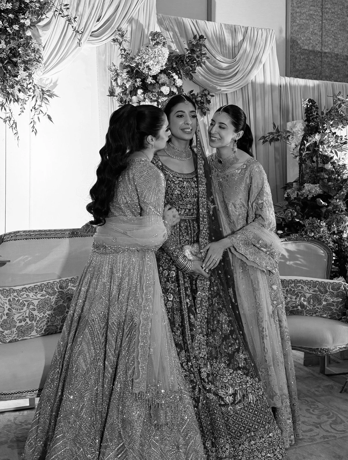 Three women in traditional Indian attire stand close together, sharing a joyful moment at a celebration venue decorated with flowers and draped fabric.