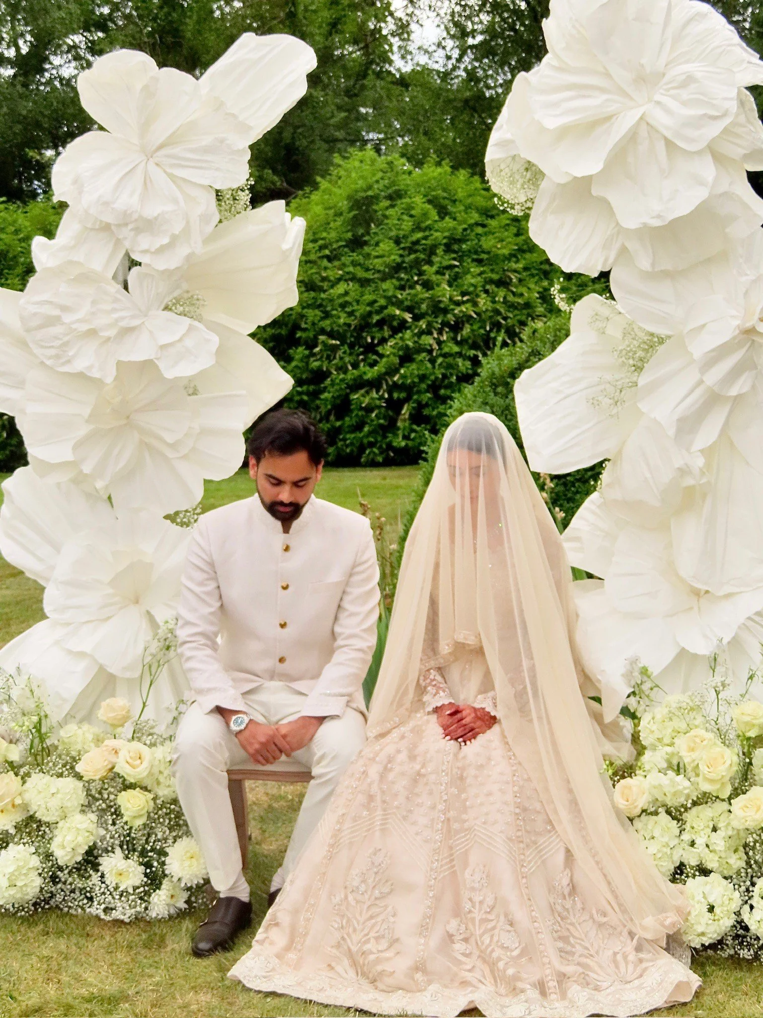 A couple during a wedding ceremony, seated in front of a floral arch with white flowers, outdoors with greenery in the background. The bride is wearing a cream-colored embroidered gown and veil, and the groom is dressed in a white suit with gold buttons.