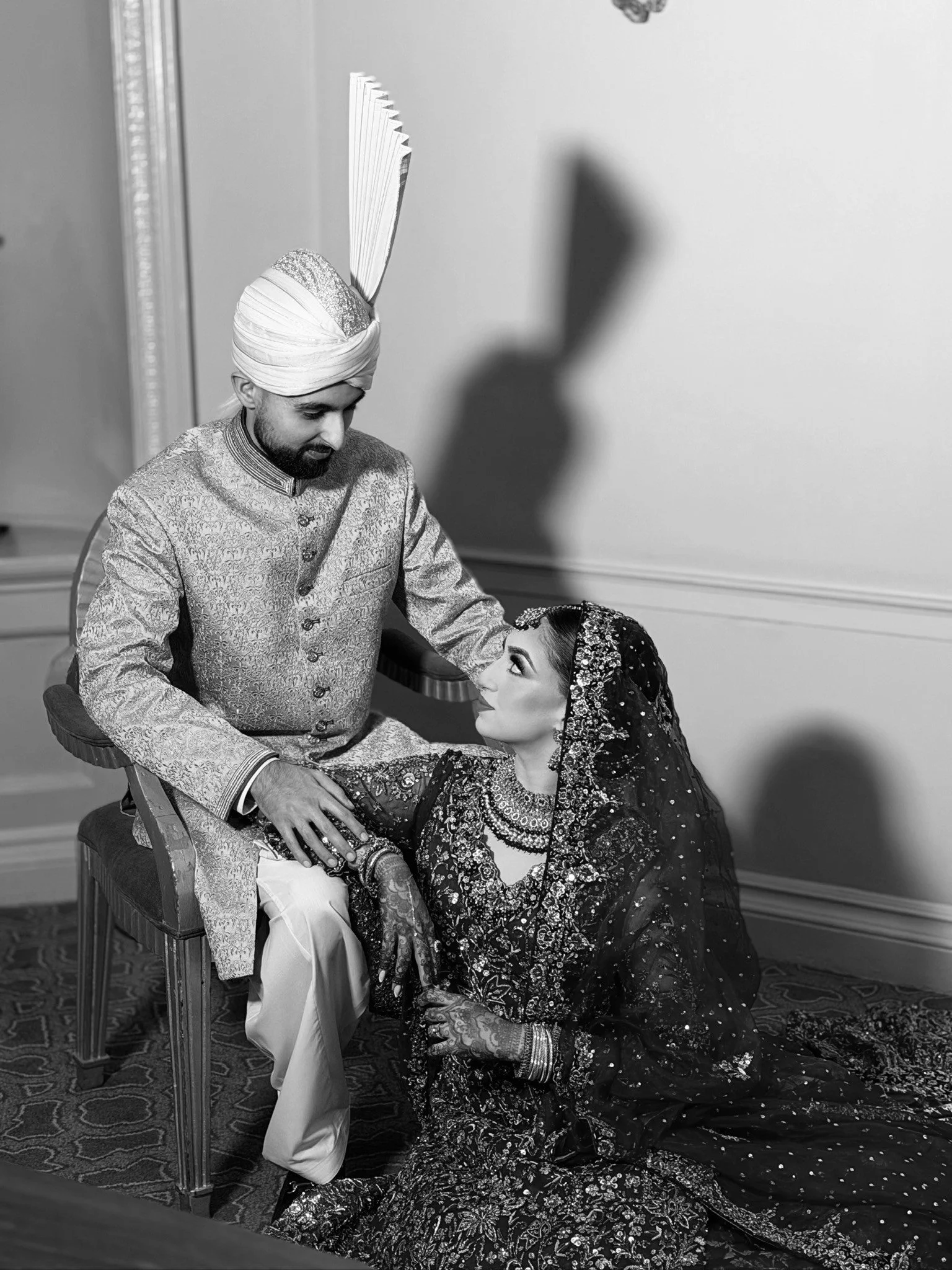 A black and white photo of a man dressed in traditional Indian attire with a turban and an ornate sherwani, sitting with a woman in traditional Indian dress, who is on the floor, looking up at him. The woman has a heavily embroidered and embellished outfit and is gazing at the man.