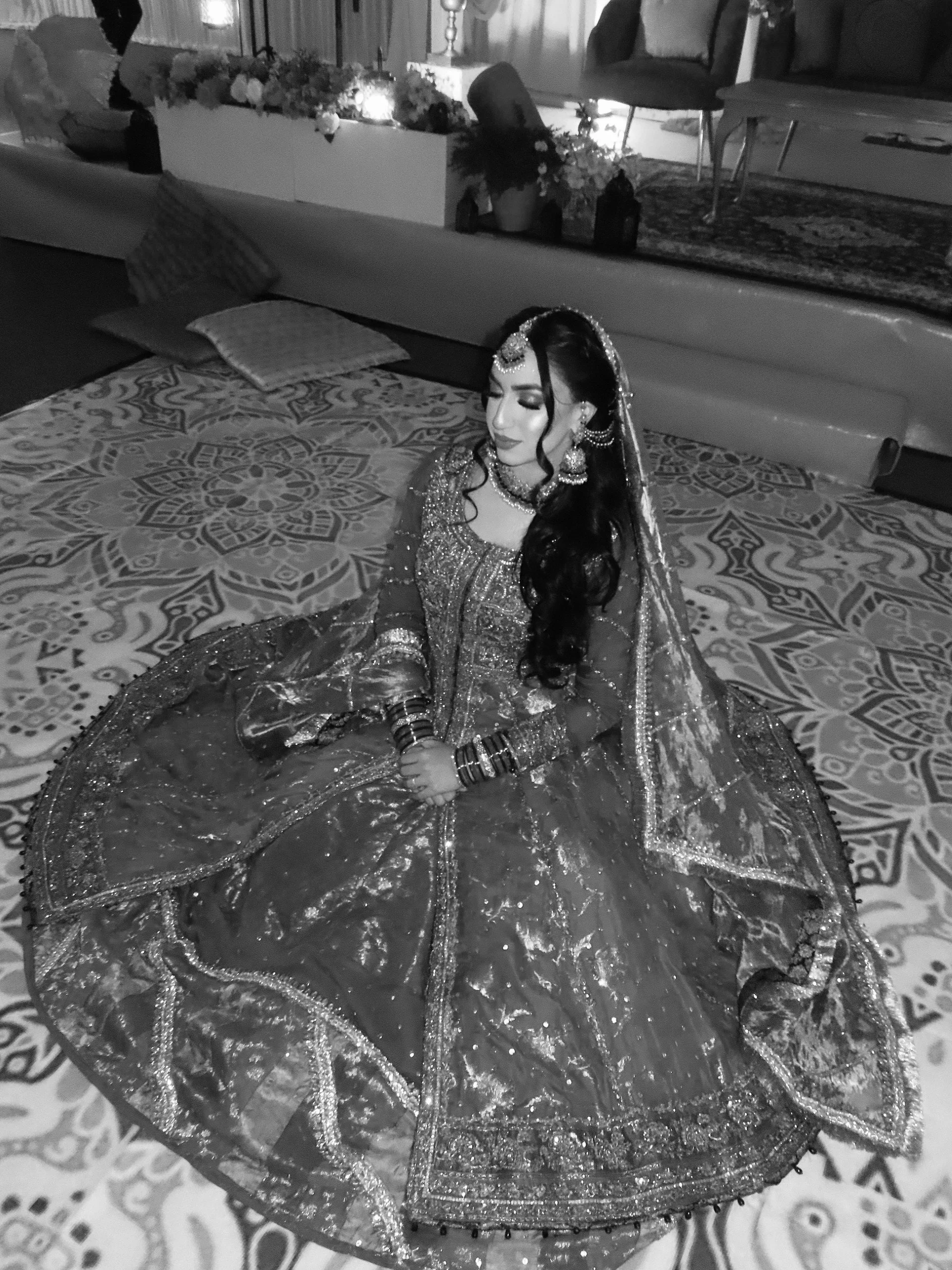 A woman dressed in traditional Indian bridal attire, sitting on a patterned carpet, with a decorated veil and jewelry.