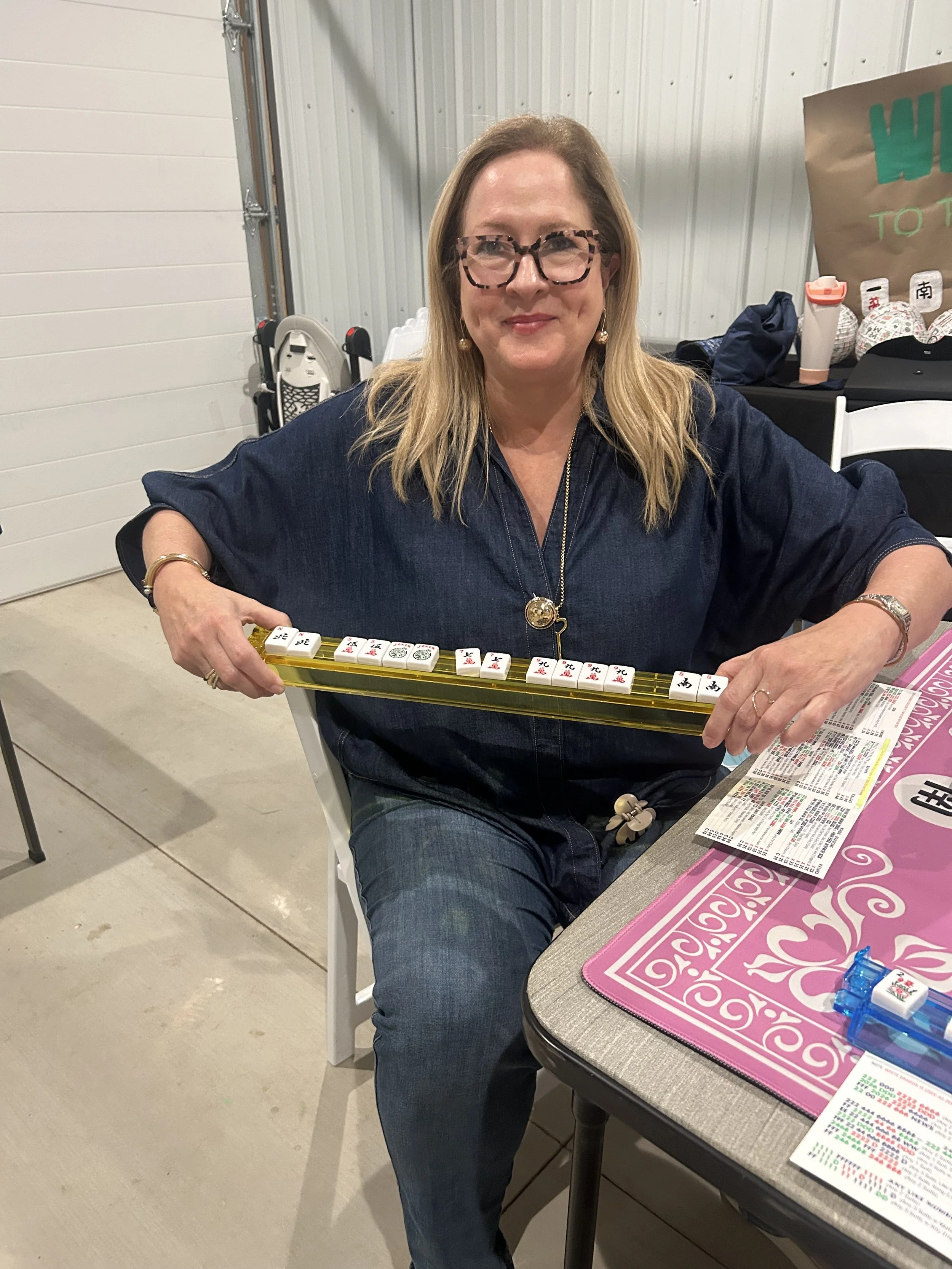 A woman with blonde hair, glasses, and earrings sitting at a table playing mahjong with Chinese character tiles. She is smiling and wearing a dark blue shirt.