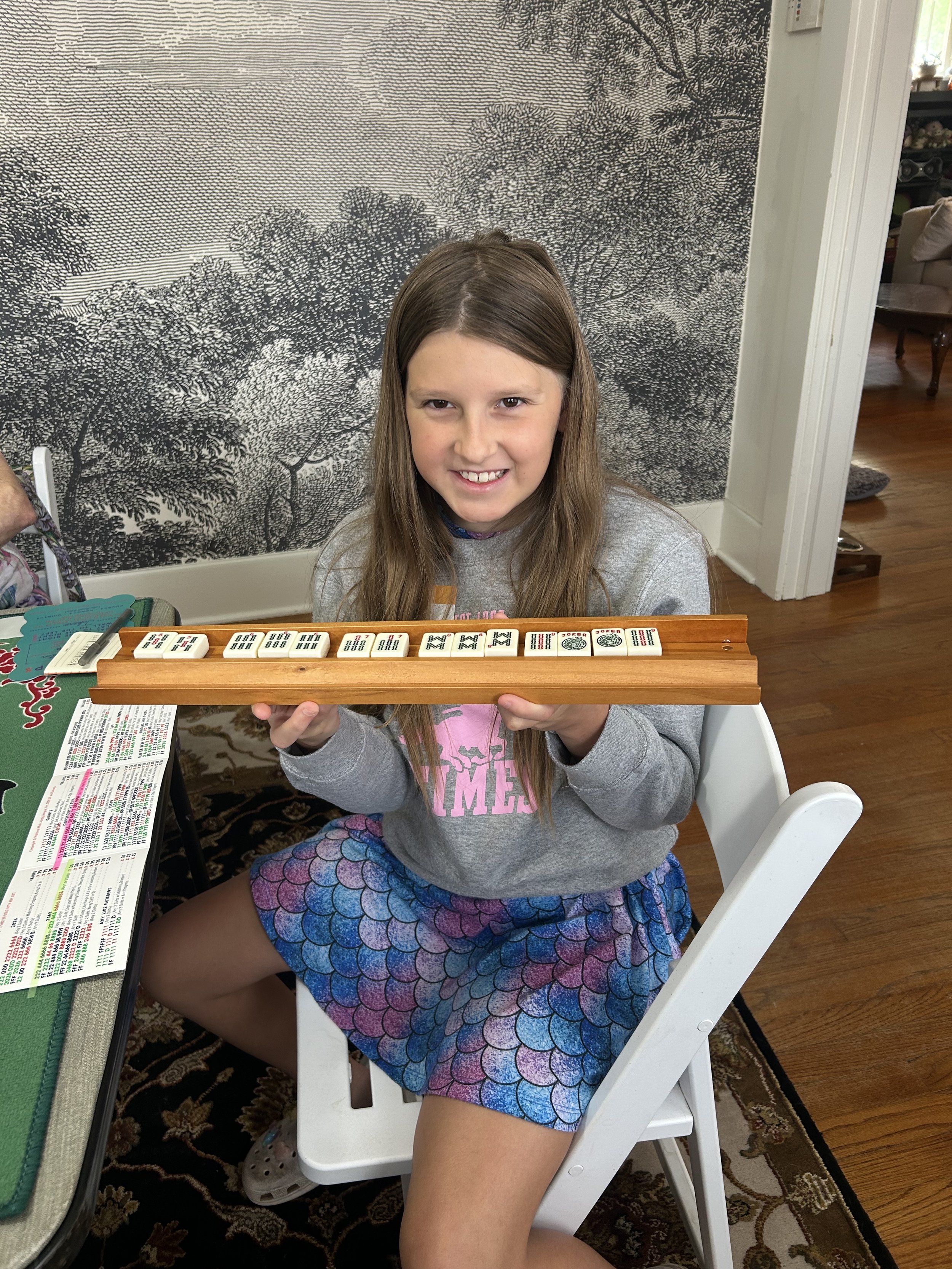 A young girl with brown hair smiling and holding a Mahjong game set indoors, sitting on a white chair. She is wearing a gray sweatshirt and colorful mermaid scale-patterned shorts.
