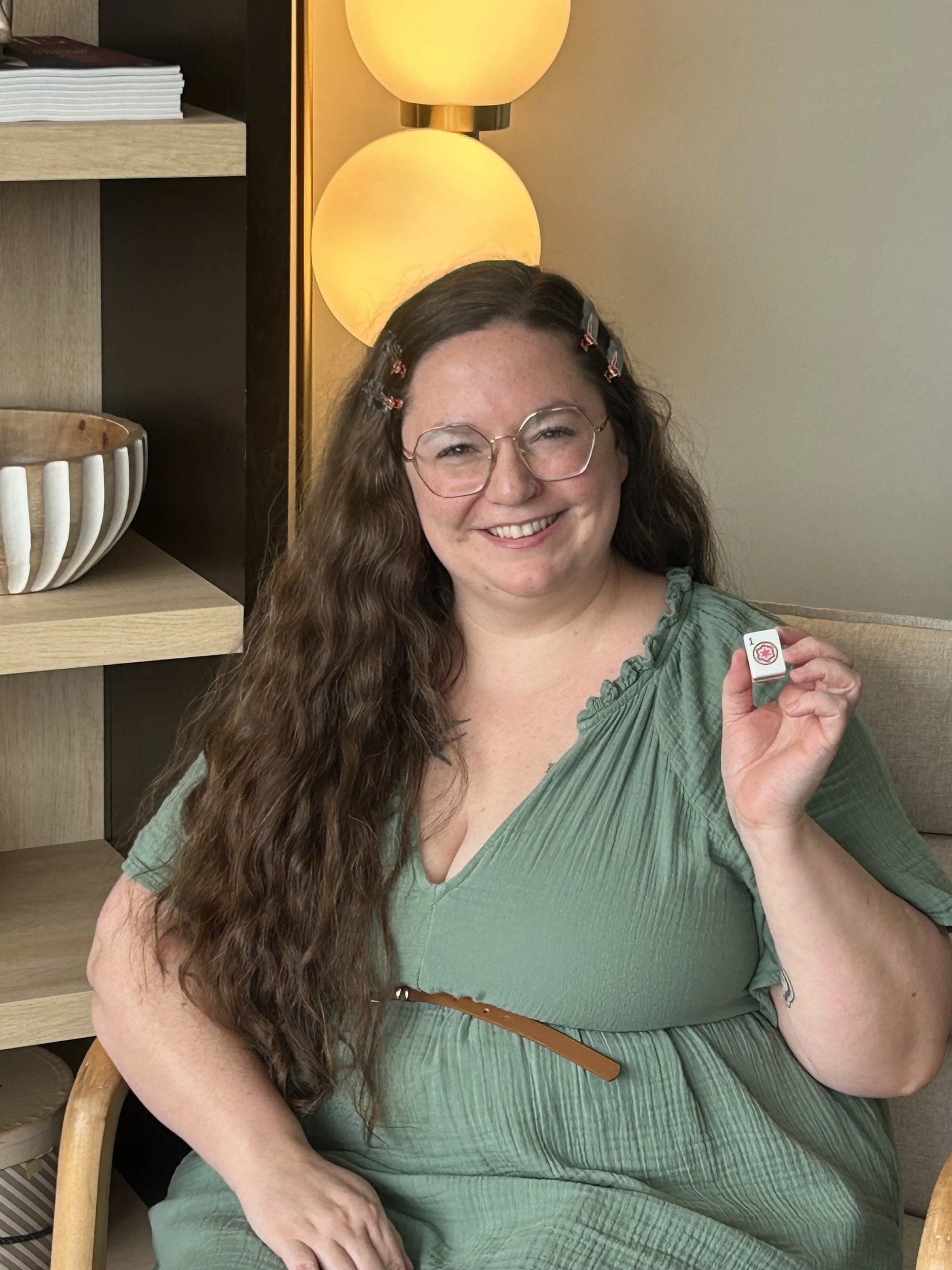 A smiling woman with long, curly brown hair, wearing glasses, a green dress, and a brown belt, seated in a room with a wooden shelf, a bowl, and a warm-colored lamp in the background, holding a small white object with a red cross.