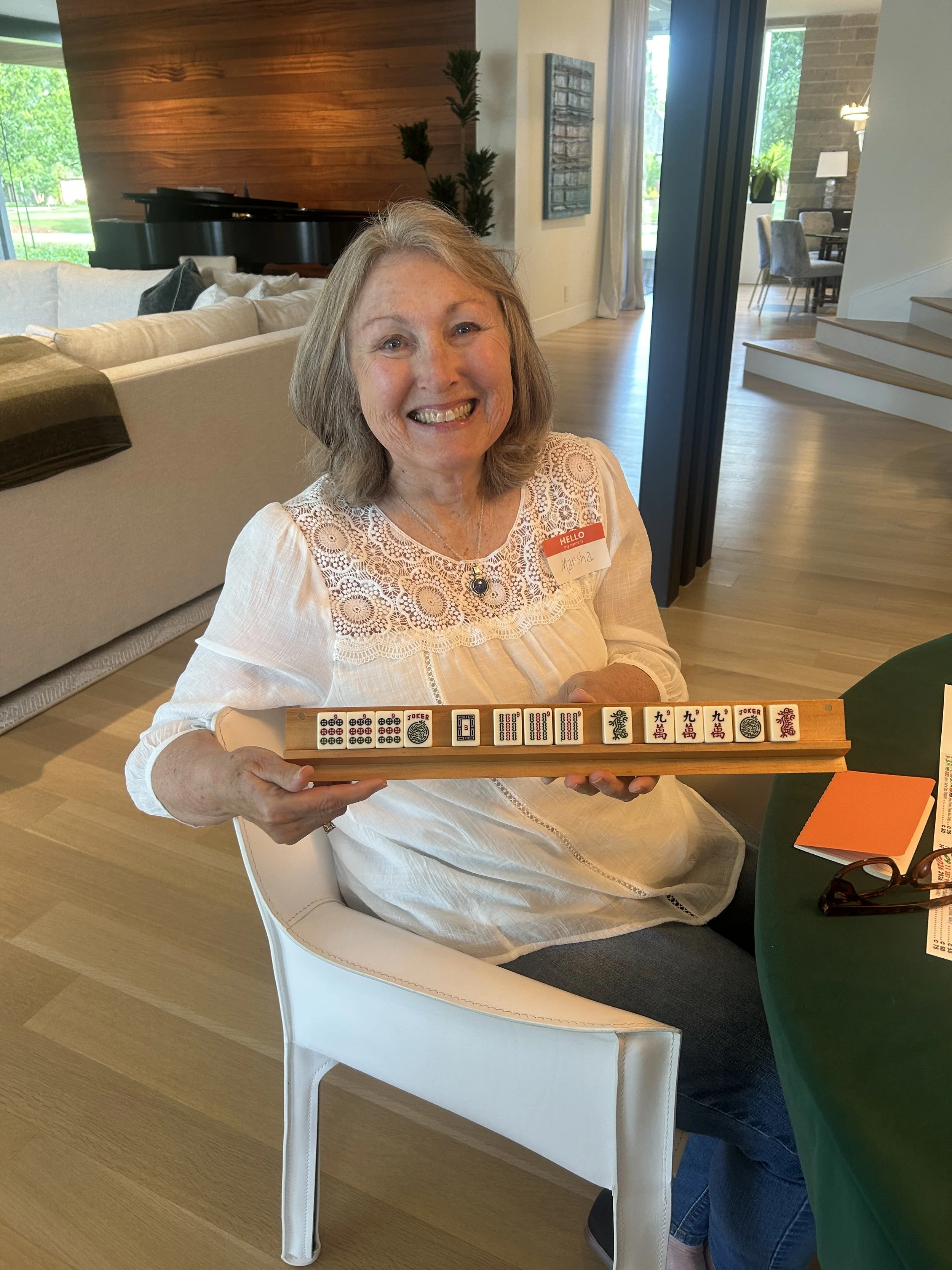 A woman with shoulder-length gray hair smiling and holding a Mahjong tile rack with tiles, sitting on a white chair at a green table in a modern living room with wooden flooring, a beige couch, and art on the walls.