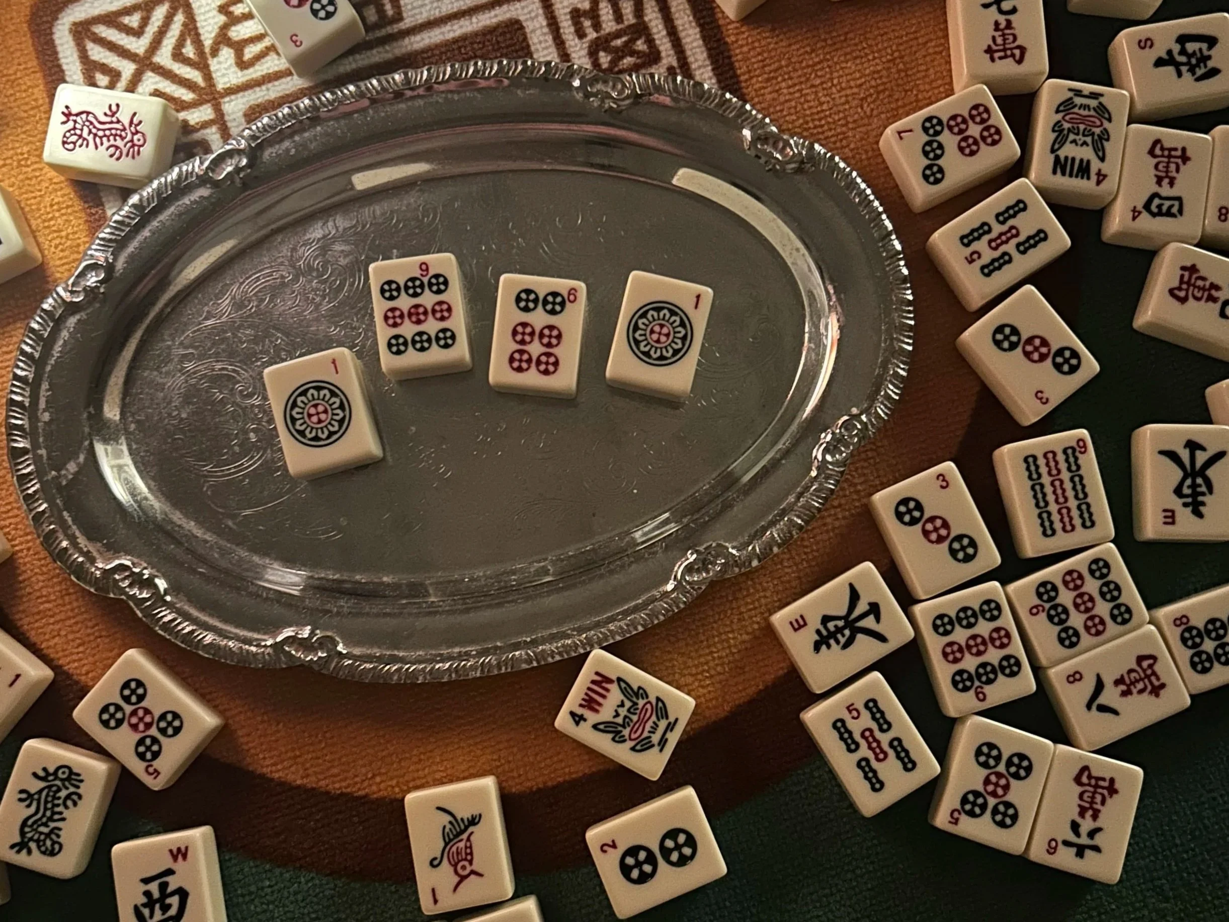 Scattered Mahjong tiles around a silver tray on a colored cloth, with some tiles on a wooden surface.