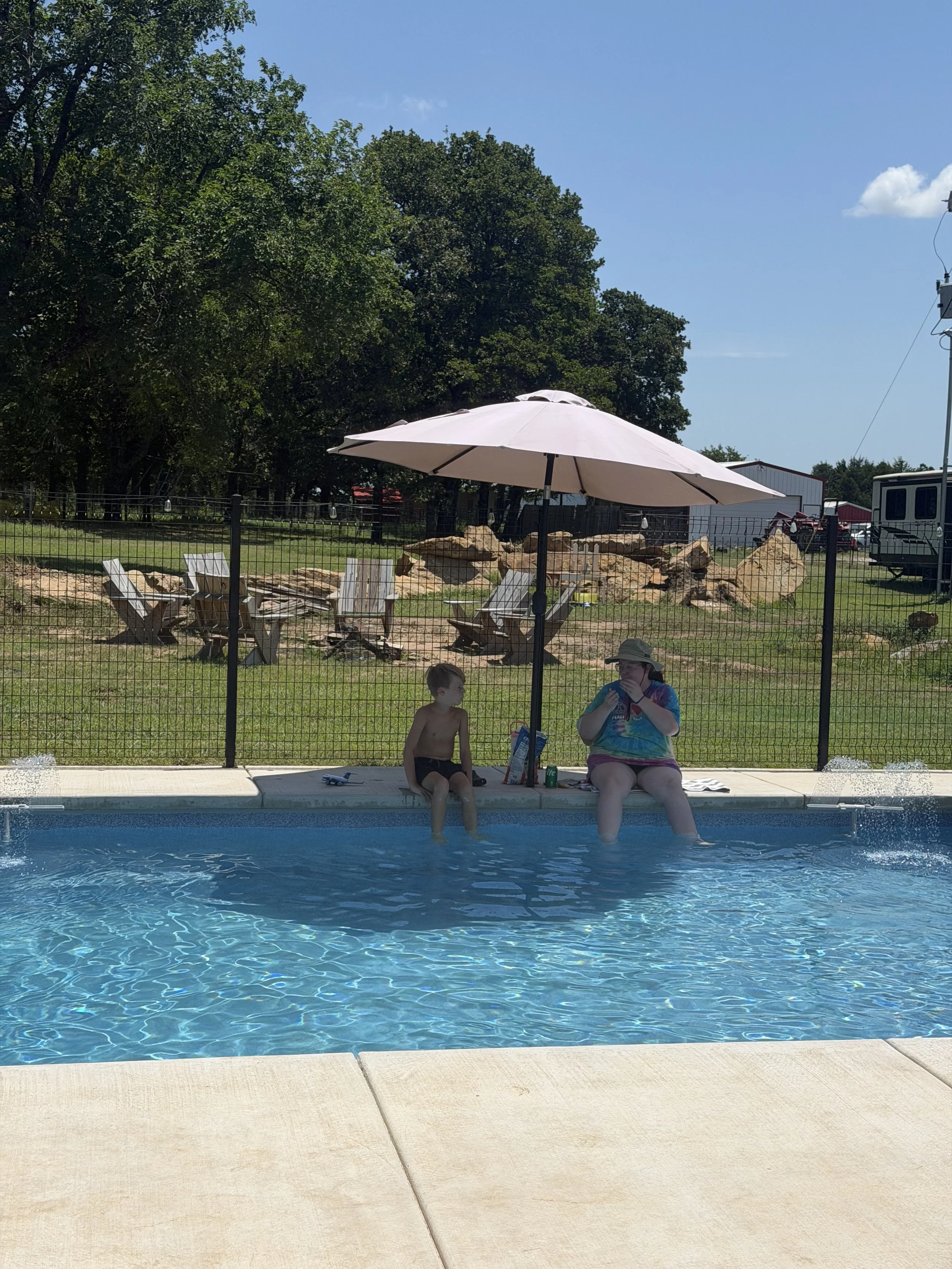 A swimming pool with two children sitting on the edge, one child is drinking from a bottle, while the other is sitting next to them. There are large rocks, a white umbrella, and green trees in the background.
