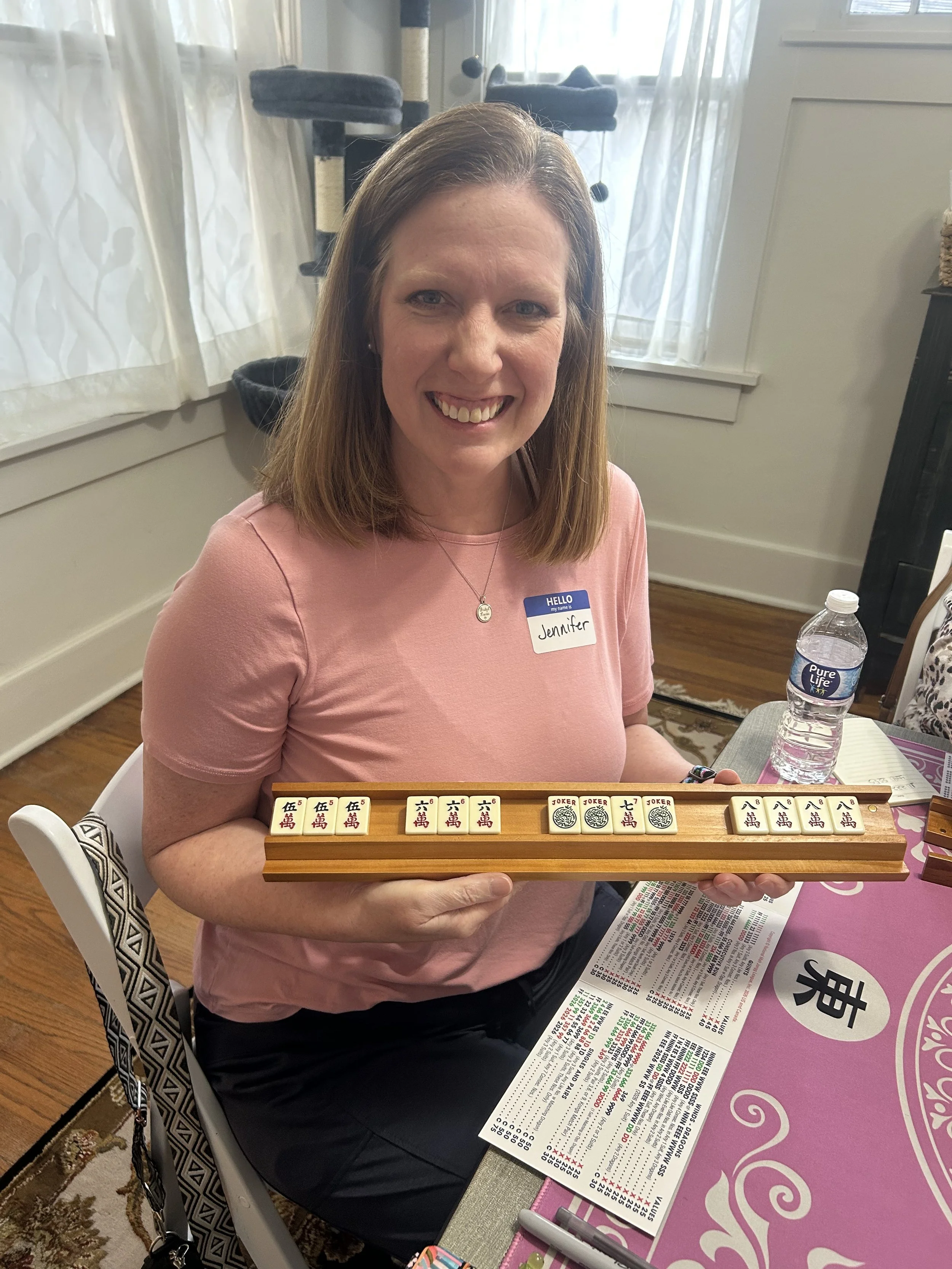 Woman smiling and holding a mahjong set at a table with a mahjong guide and a water bottle, wearing a pink shirt with a name badge that says 'Jennifer'.