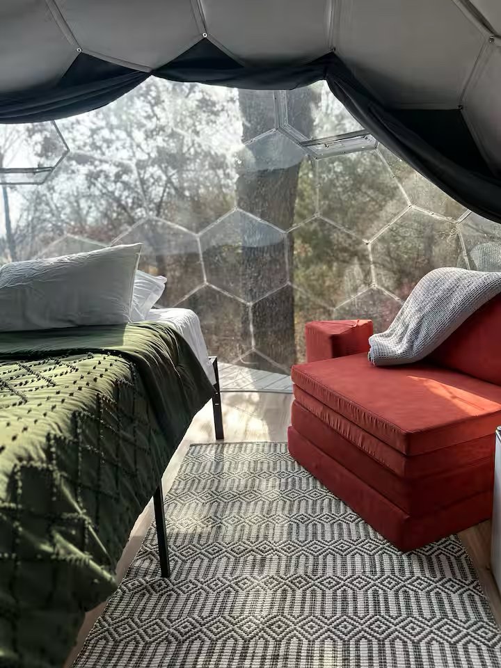 Bedroom with a bed on the left, a red armchair on the right, and a large transparent hexagonal window showing outdoor trees and sunlight.