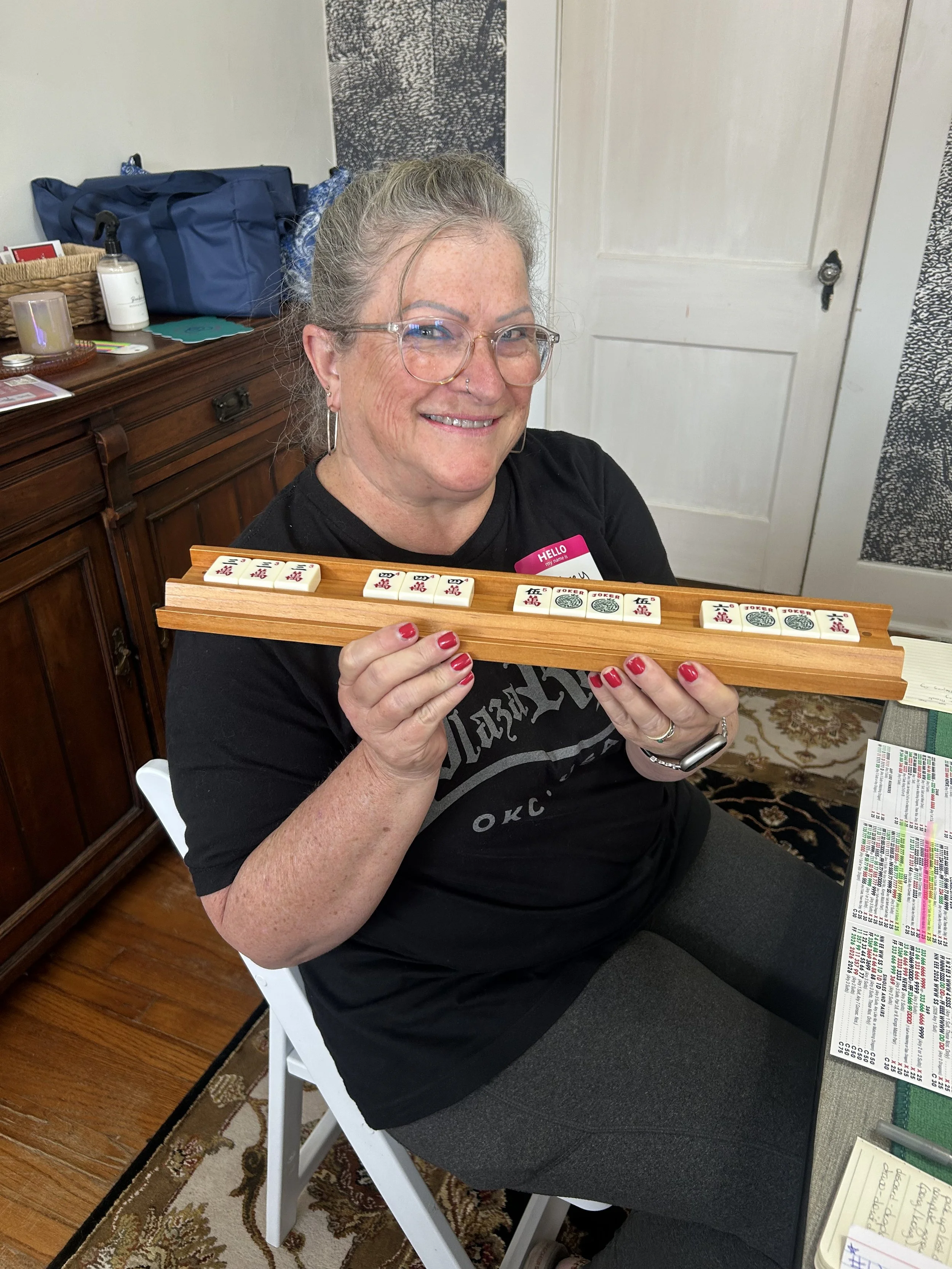 A woman with gray hair, glasses, and a nose piercing smiling and holding a wooden Mahjong tile rack with Mahjong tiles on it, sitting at a table with papers and a pen, in a room with wooden flooring, a wooden sideboard, and a decorative rug.