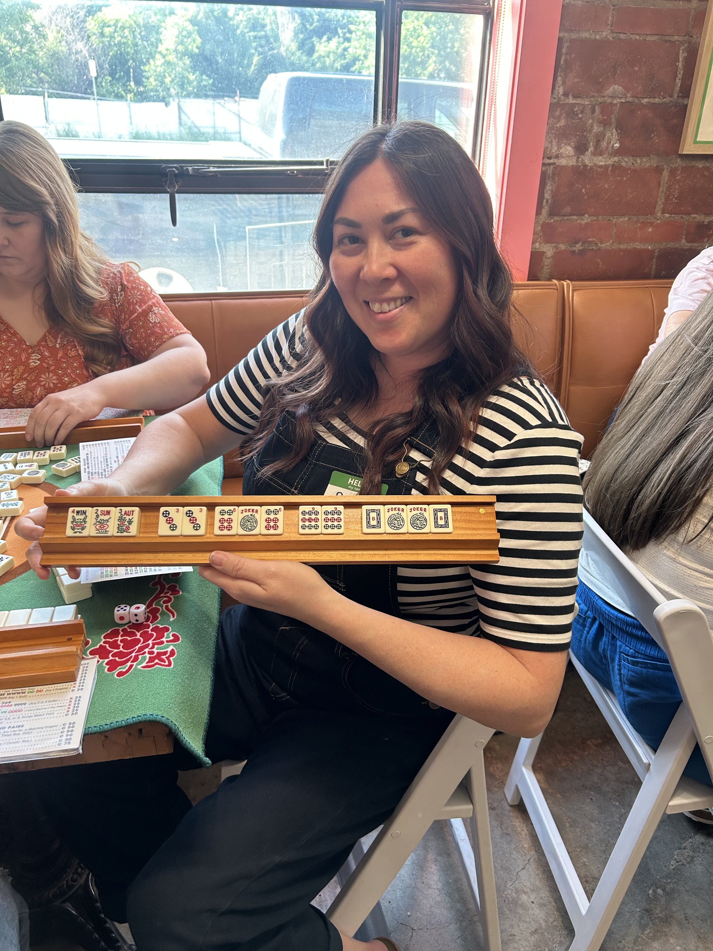 A woman smiling and holding a wooden domino set, sitting at a green table with other people playing dominoes in a restaurant with brick walls and large window.