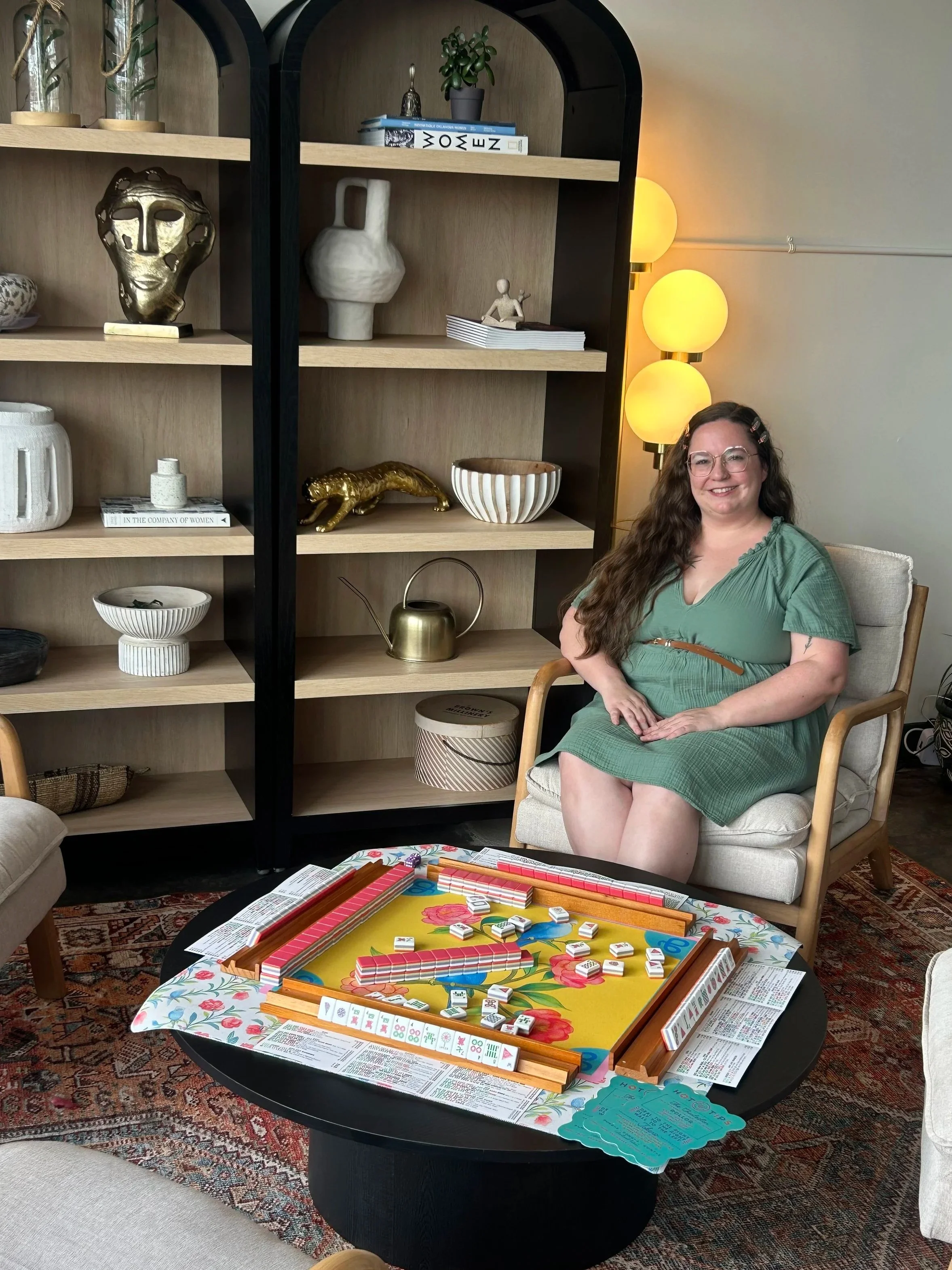 A woman with glasses and long brown hair sitting on a cream-colored armchair in a living room. Behind her is a black bookshelf with decorative items, including a gold face sculpture, white vases, a small sculpture, and books. To the right of the woman, there is a floor lamp with three round yellow globes. In the foreground, a round table with a colorful floral tablecloth holds a board game with domino tiles, a sheet with printed text, and some cards.